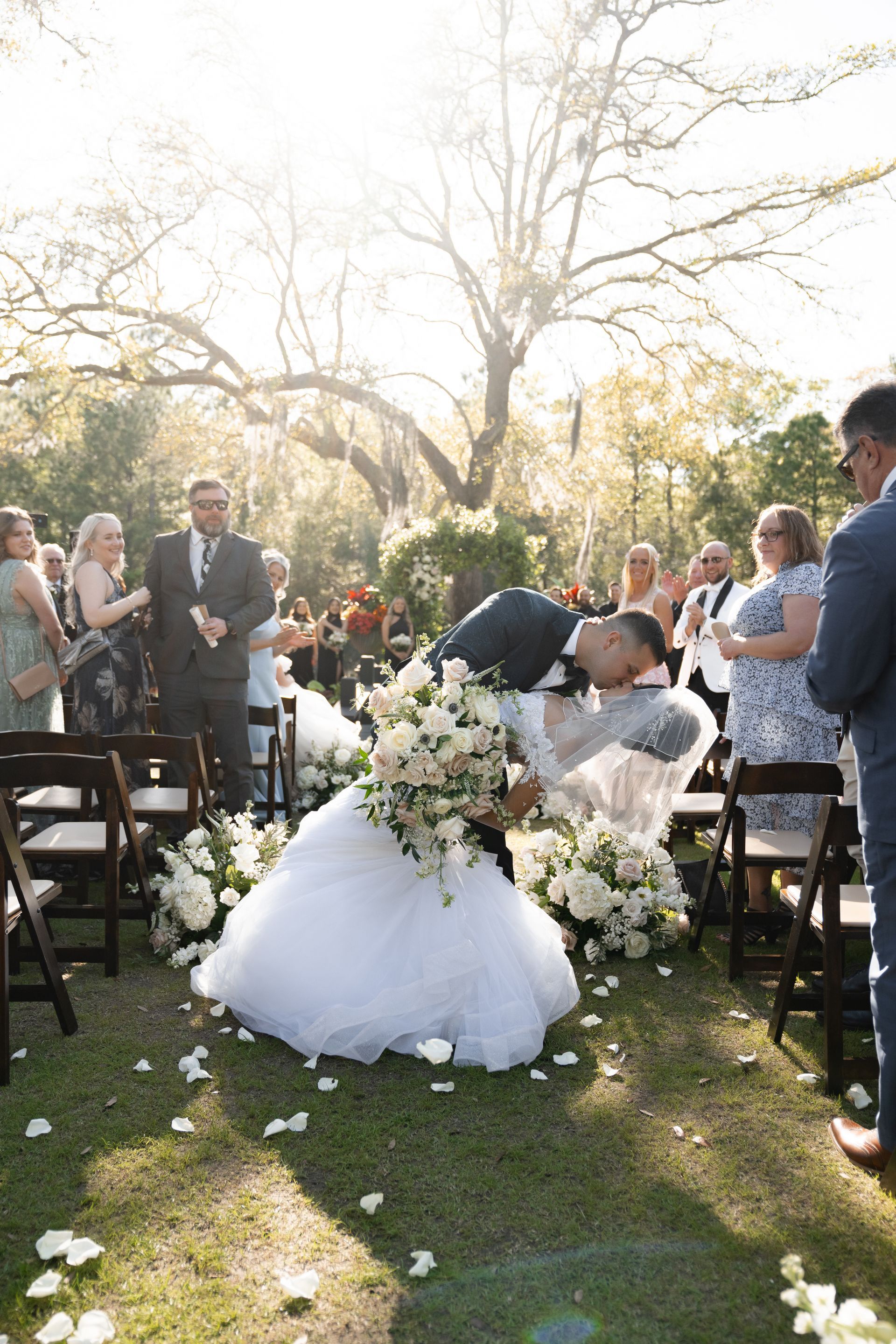 Bride and groom kiss at an outdoor wedding aisle surrounded by guests and white flowers