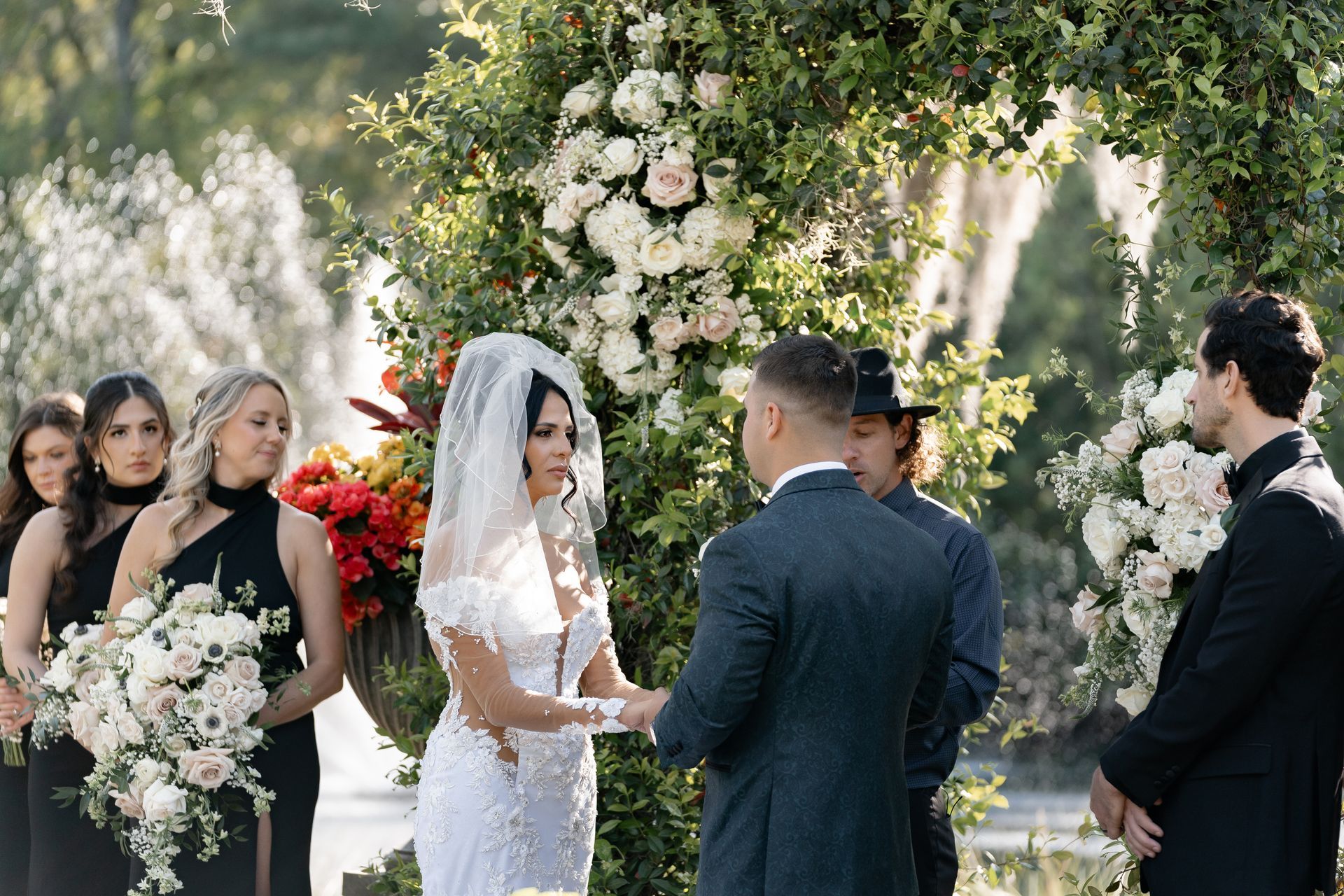 Wedding ceremony outdoors under a floral arch, bride in white facing groom in navy suit, guests nearby
