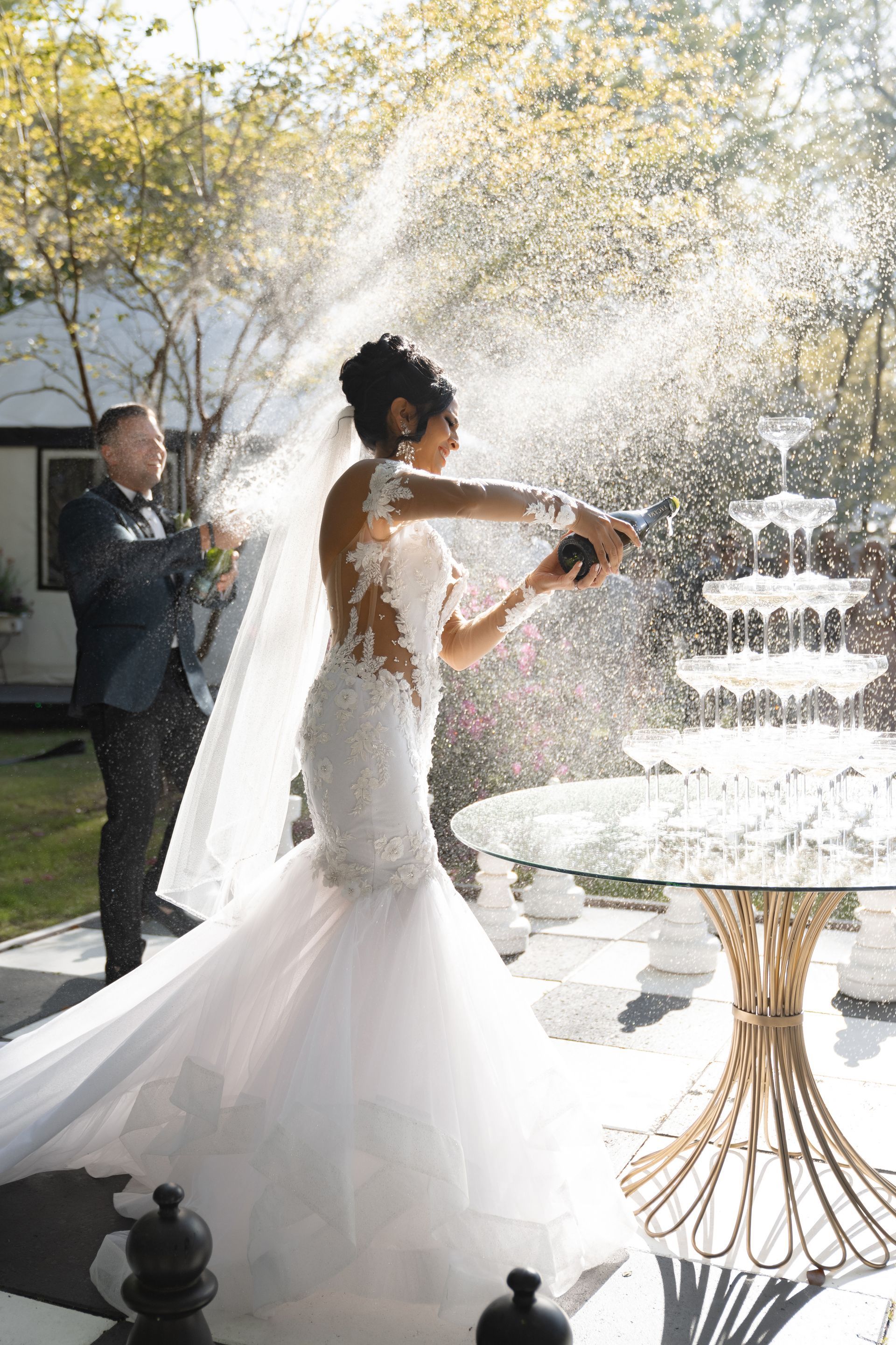 Bride in a white gown sprays champagne outdoors at a fountain, with a guest watching nearby.