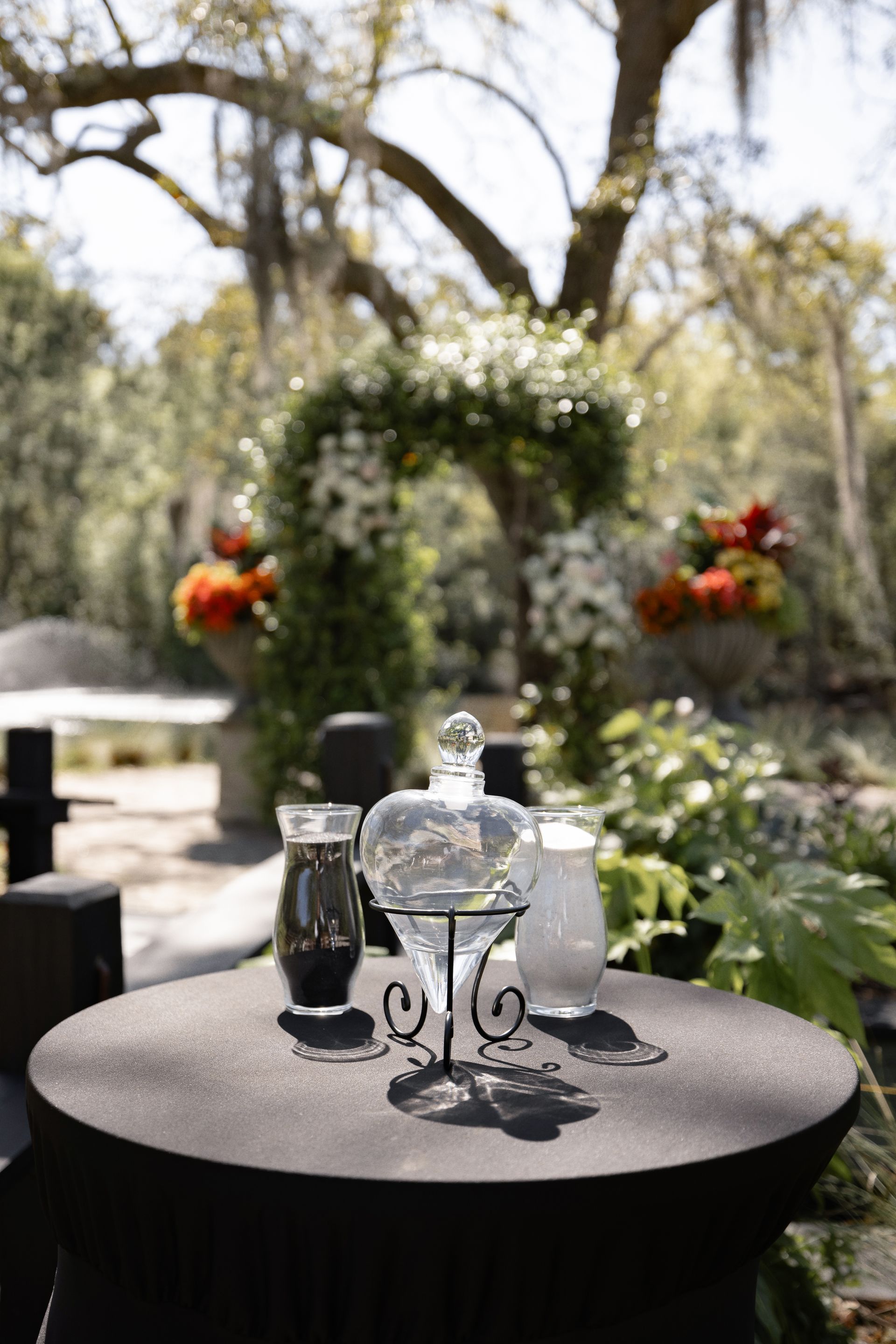Outdoor table setting with glass pitcher and jars on a black table, under a floral arch in a garden.
