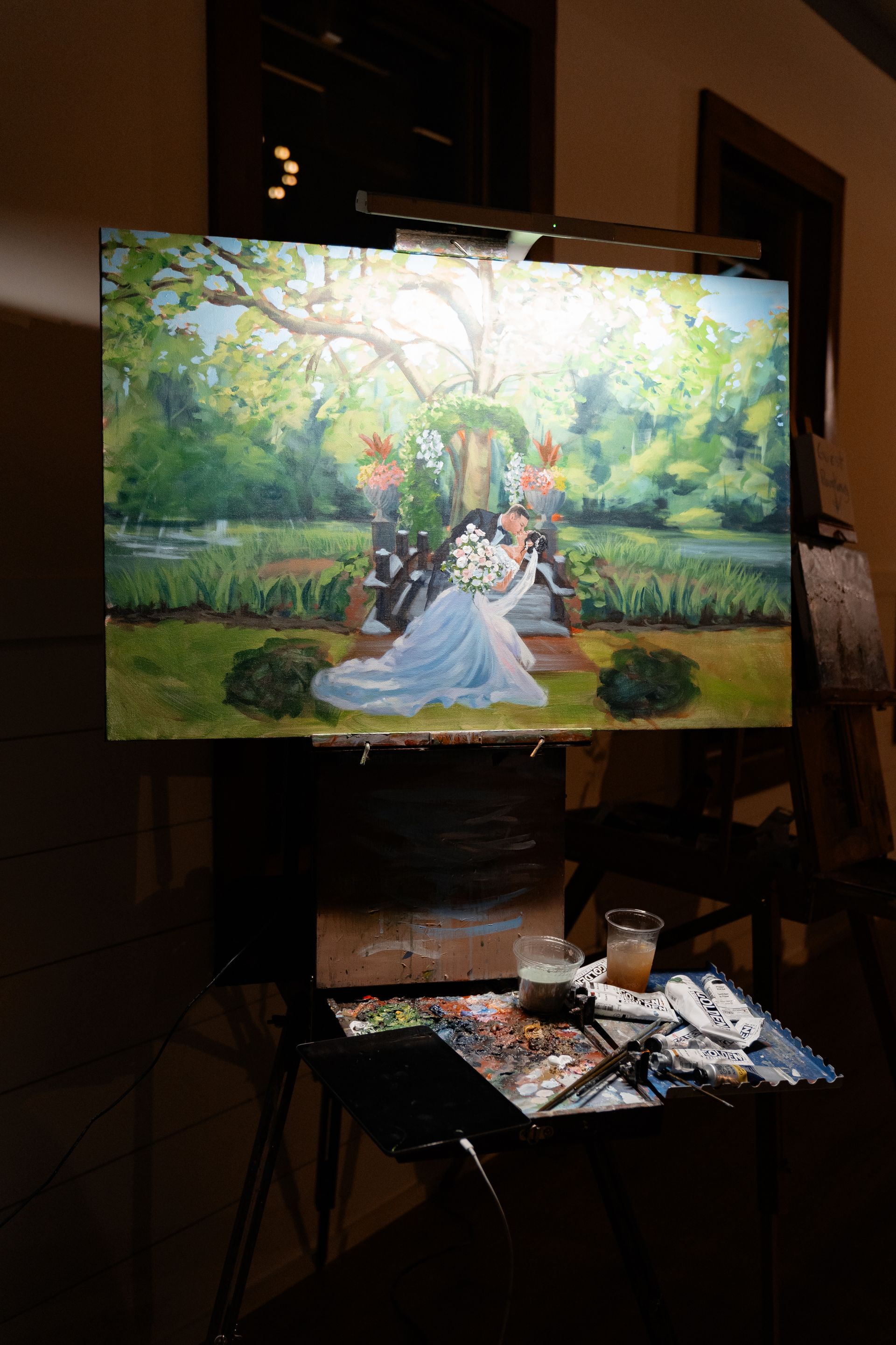 Artist painting a wedding scene on an easel in a dim studio, with paint supplies on a table.