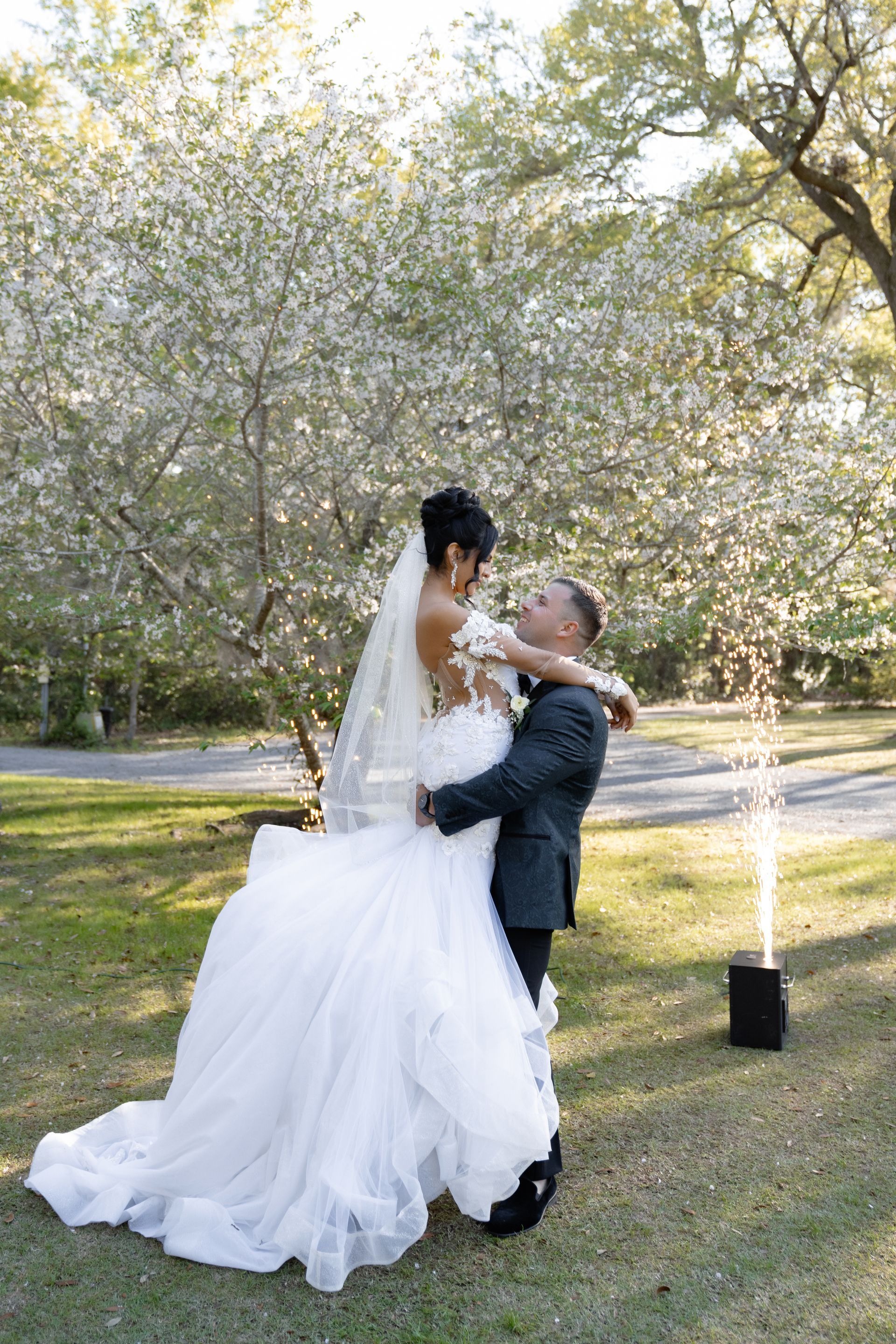 Bride in a white gown and groom in a dark suit embracing outdoors under blooming trees.