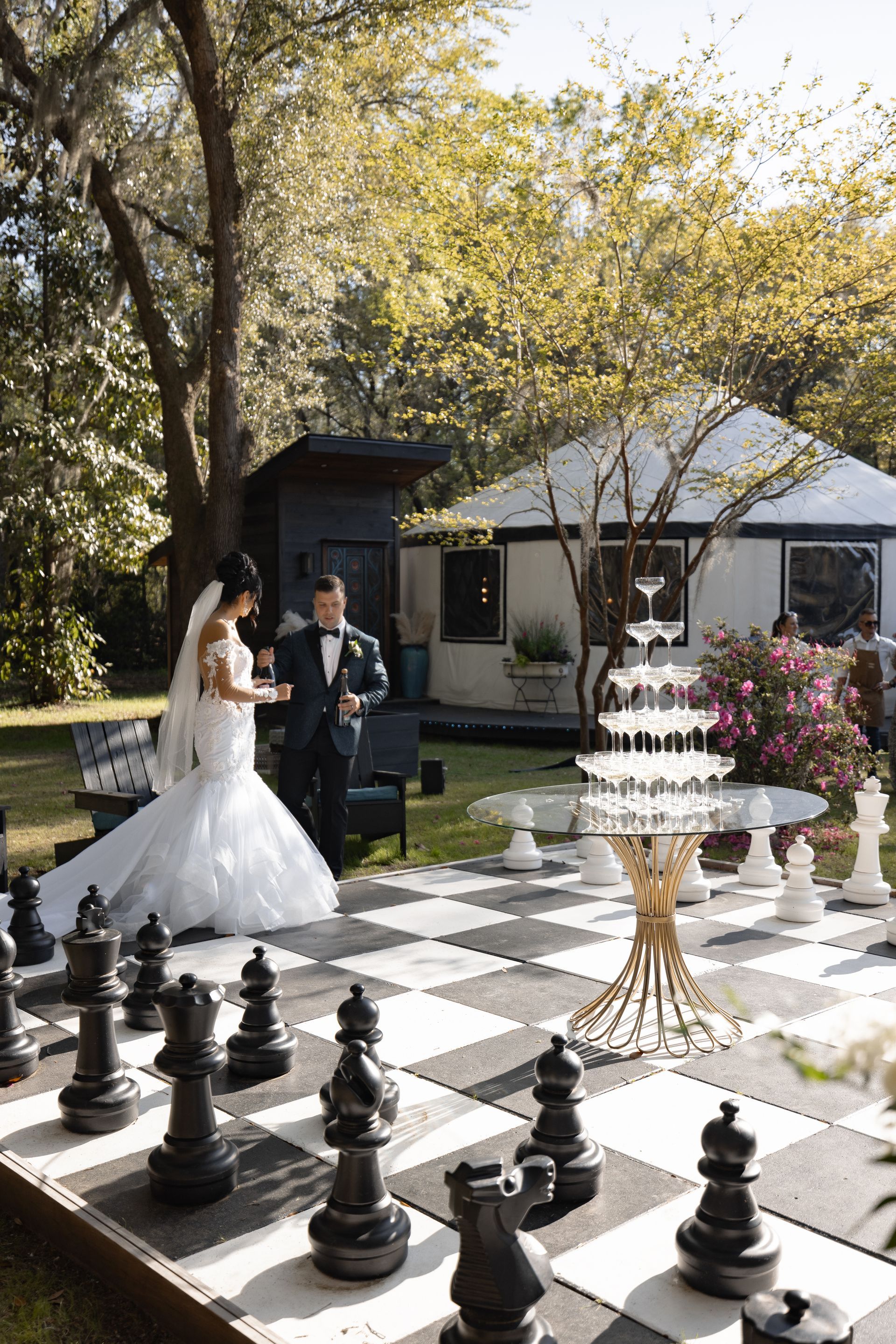 Bride and groom stand on giant chessboard at outdoor wedding reception with white tent and trees
