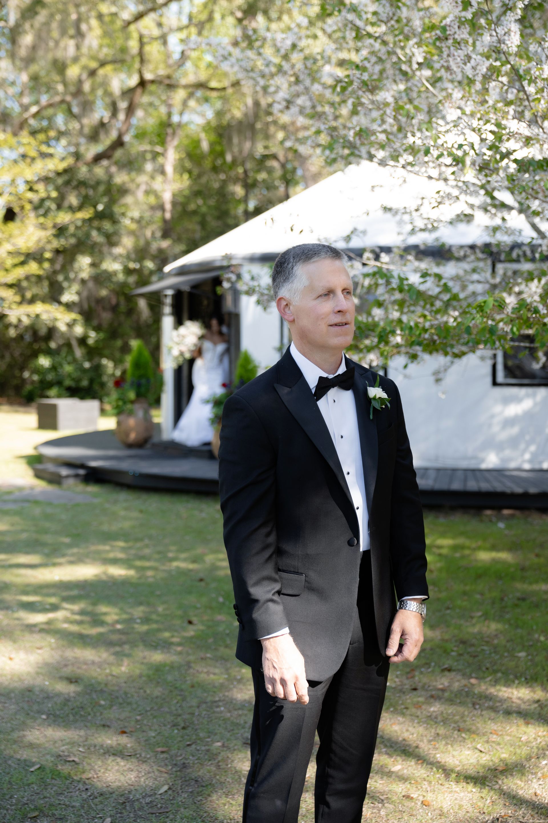 Man in a black tuxedo standing on grass near a white tent in a sunny outdoor setting