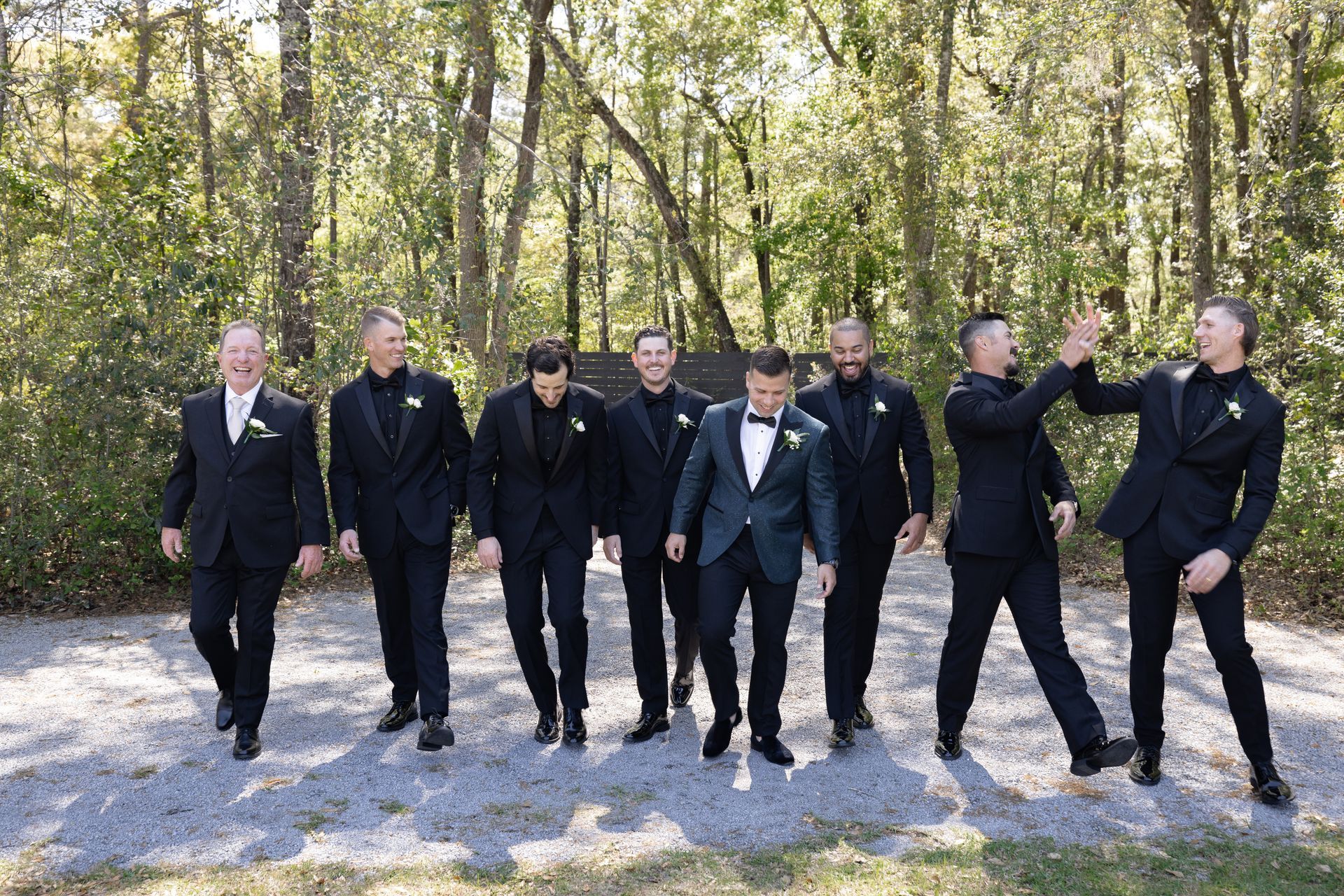 Eight people in black suits walking together on a wooded path in daylight.
