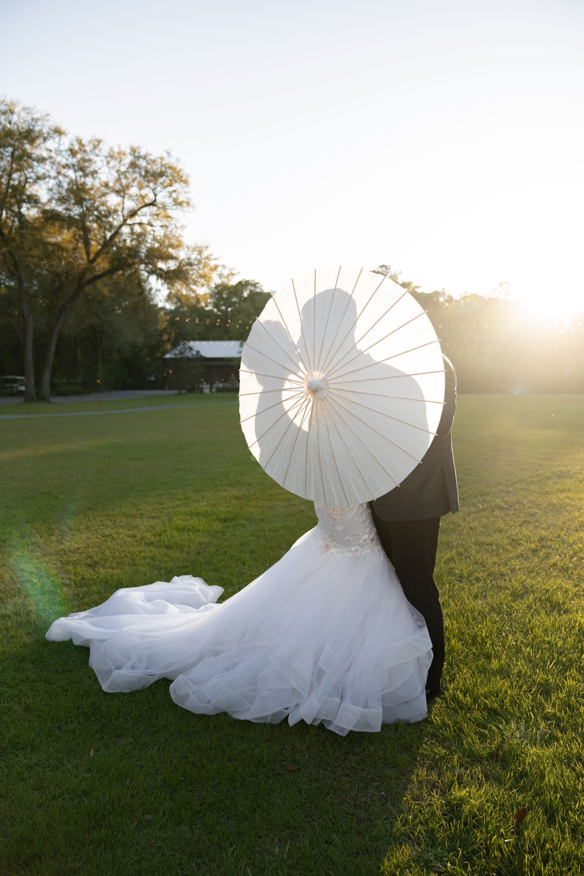 Bride in a white gown holding a white parasol on a sunlit grassy field at sunset