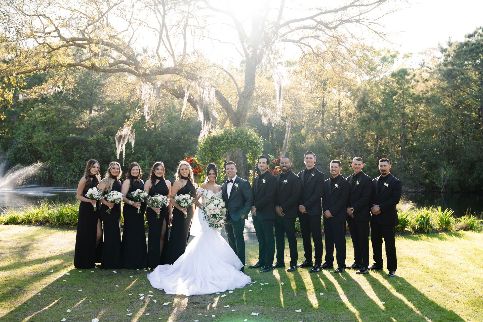 Wedding party posing outdoors in formal black and white attire under sunlit trees