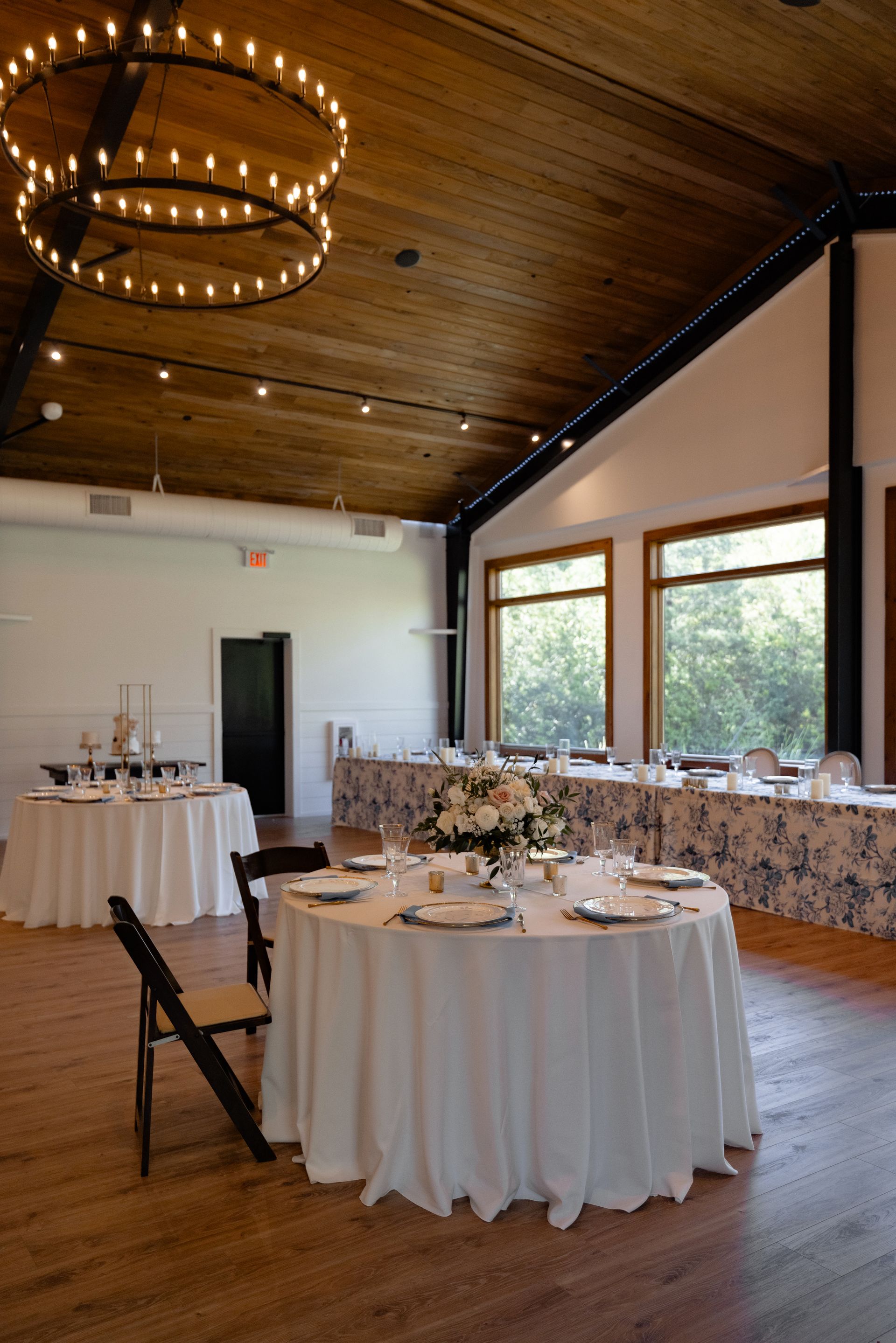 Elegant banquet hall with round tables, white tablecloths, floral centerpieces, and large windows under a wood ceiling