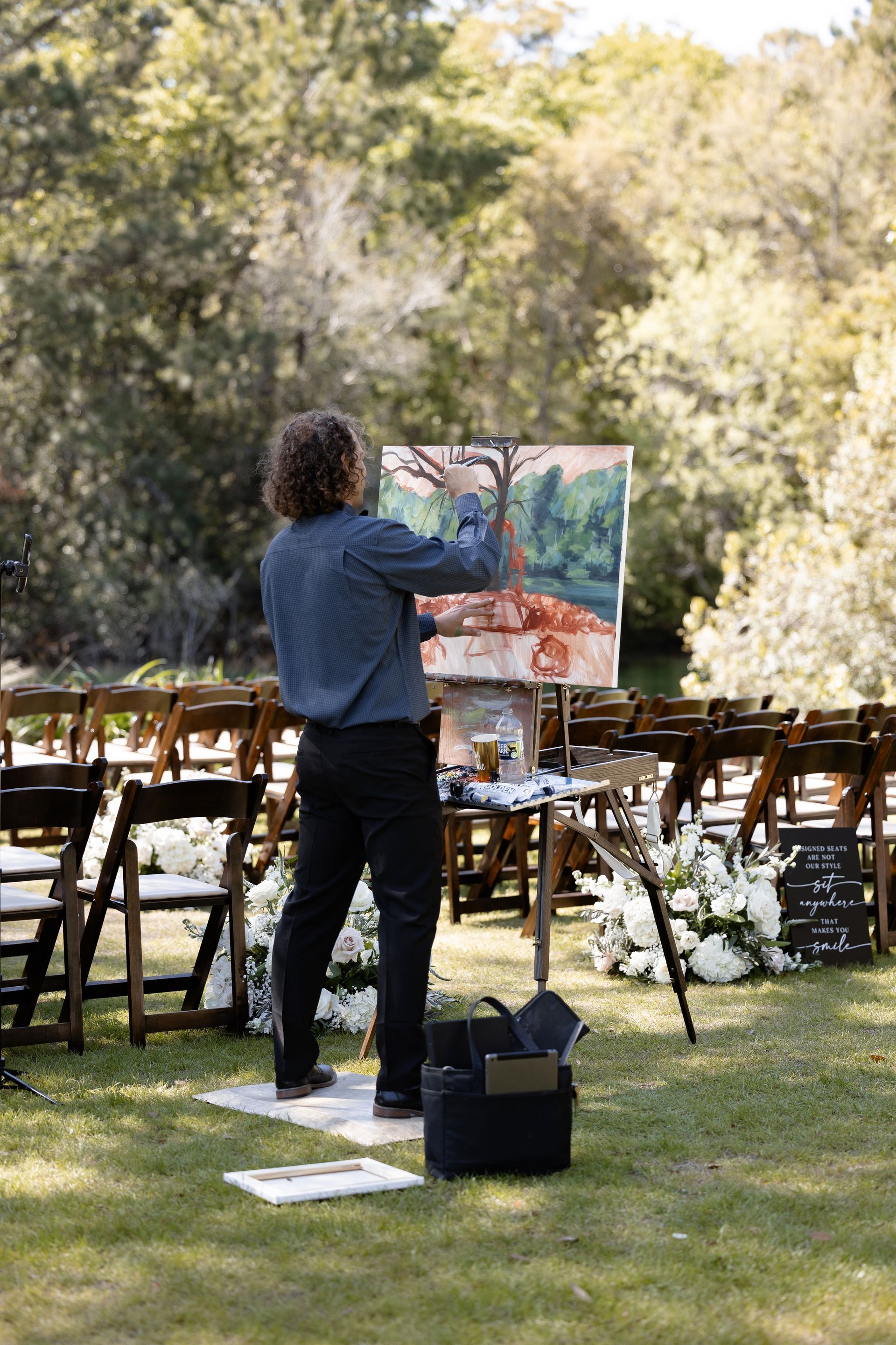 Artist painting outdoors at an easel during an outdoor wedding setup, with chairs and floral arrangements nearby