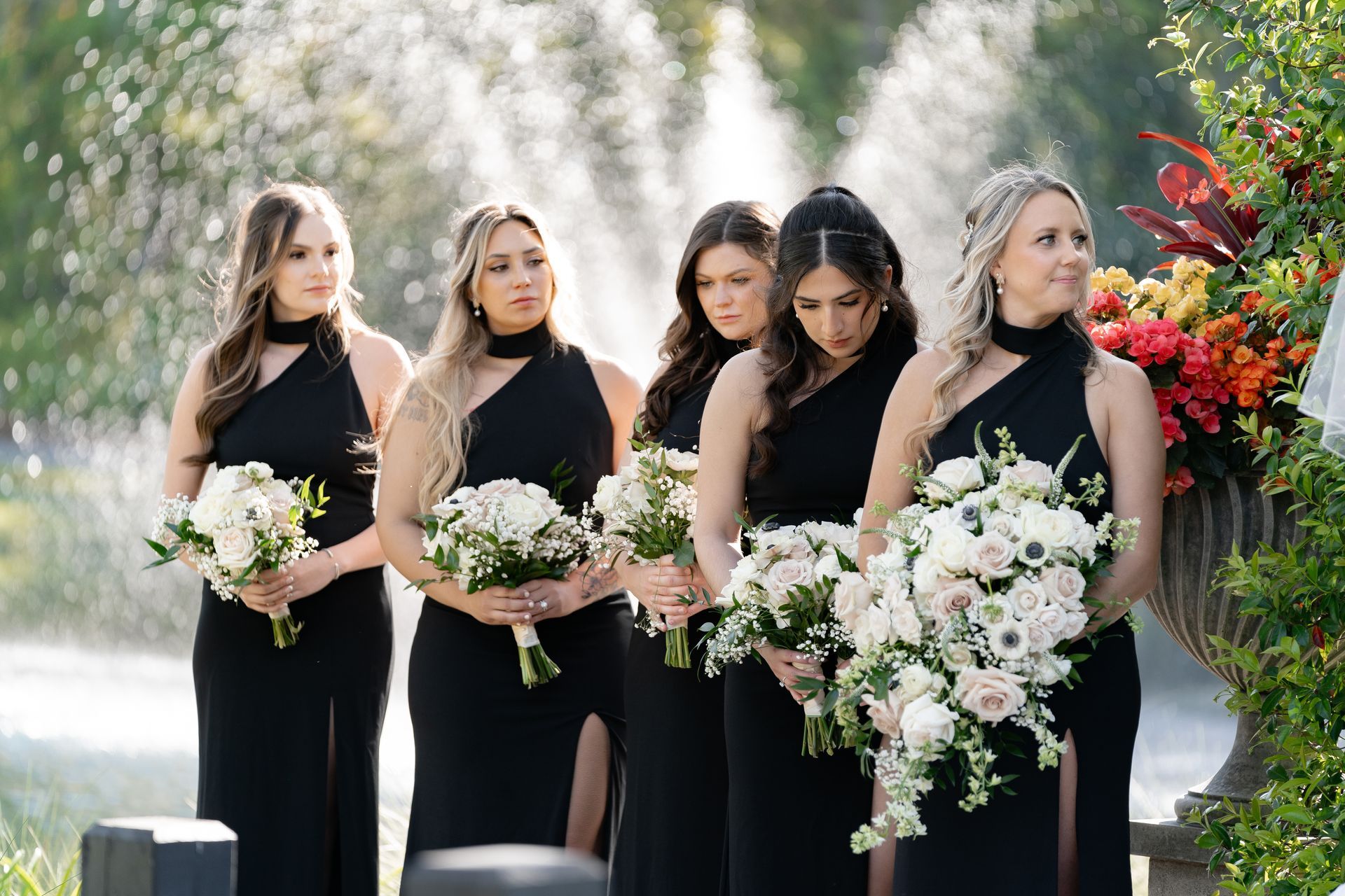 Five women in black dresses holding white flower bouquets outdoors in a wedding procession