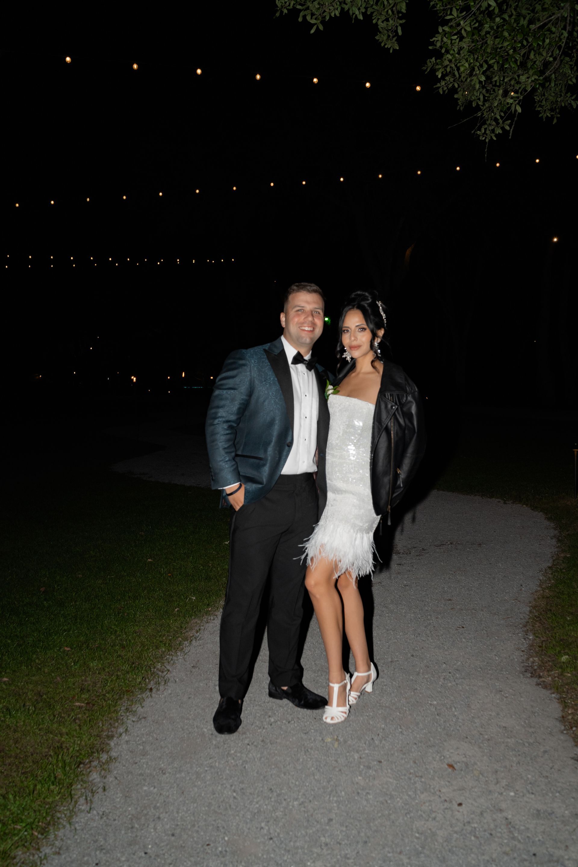 A couple posing outdoors at night on a gravel path, with string lights in the background.