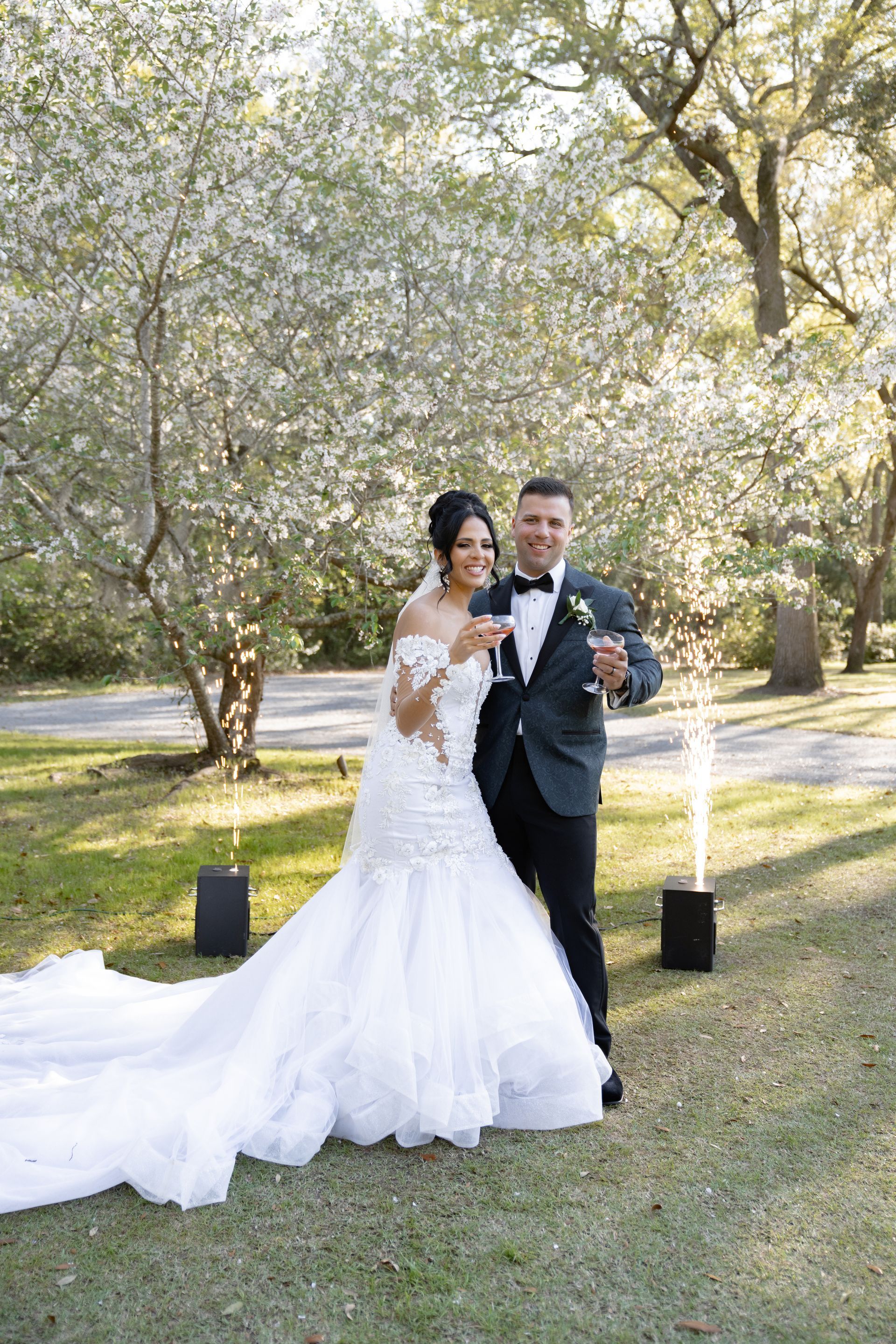 Bride and groom posing outdoors under blooming trees, bride in white gown and groom in black tuxedo