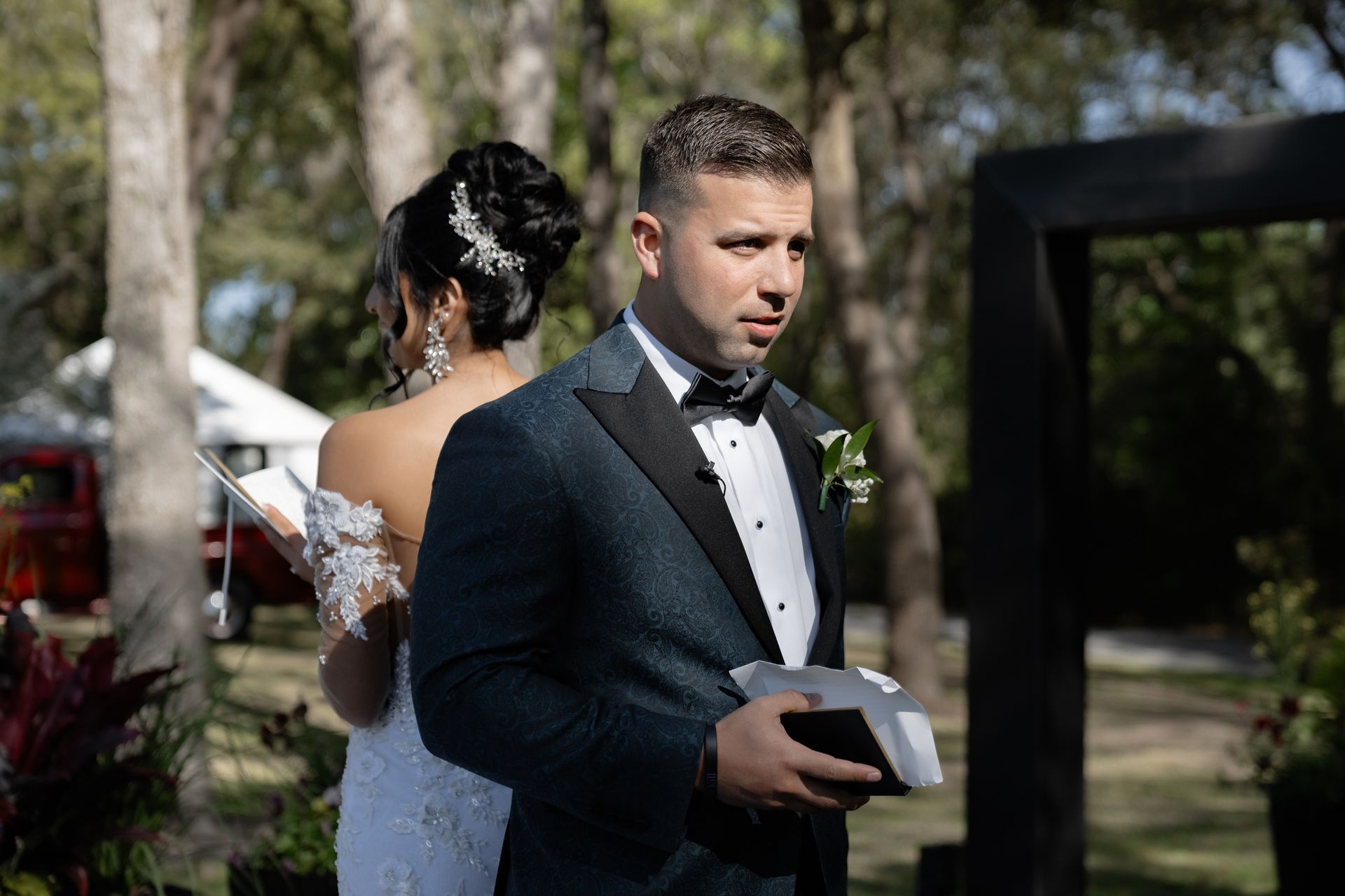 Man in a navy tuxedo holding a book, standing beside a woman in a white dress outdoors at a wedding ceremony