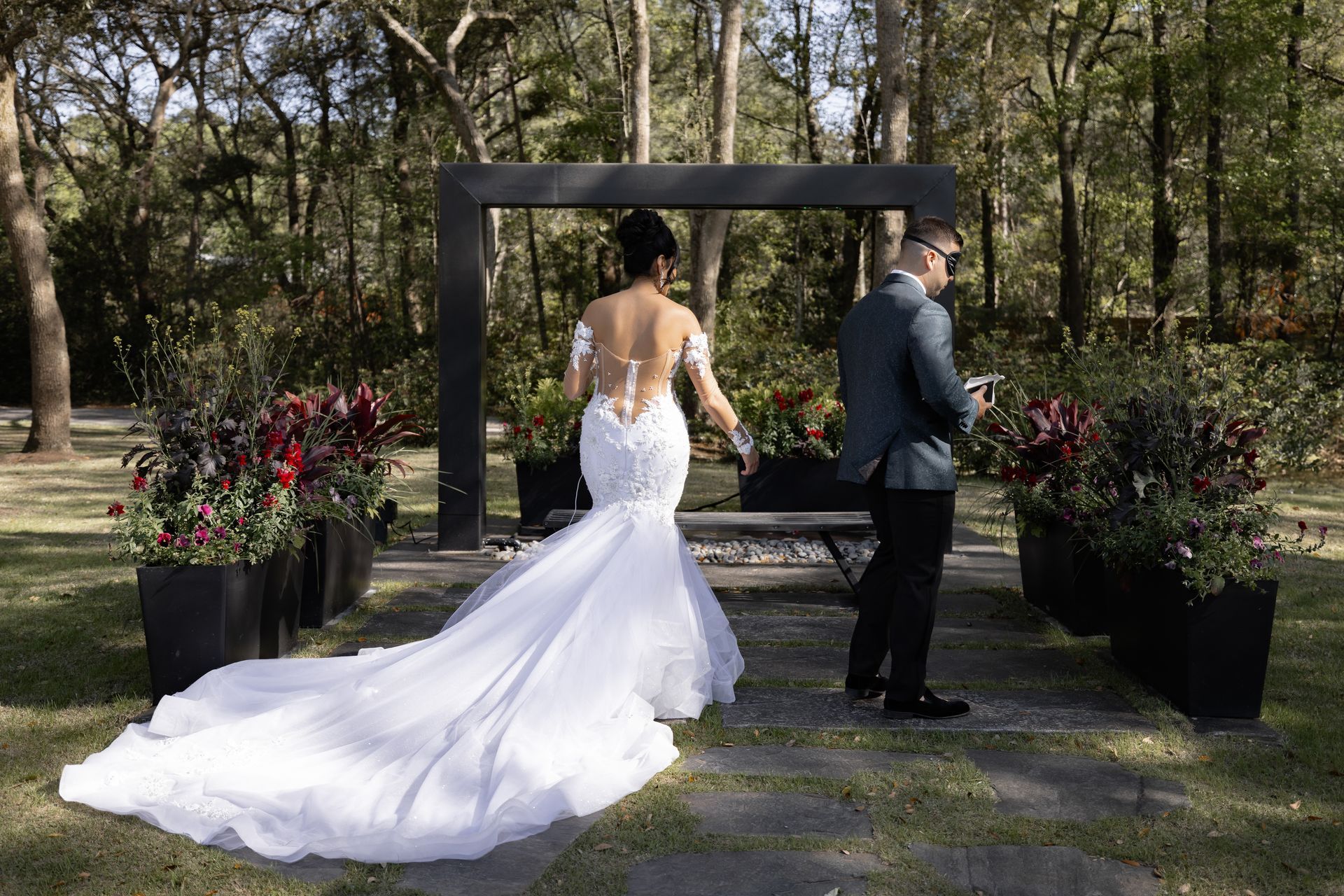 Bride in a white gown and groom in a suit walk toward a wooden arch in a wooded garden.
