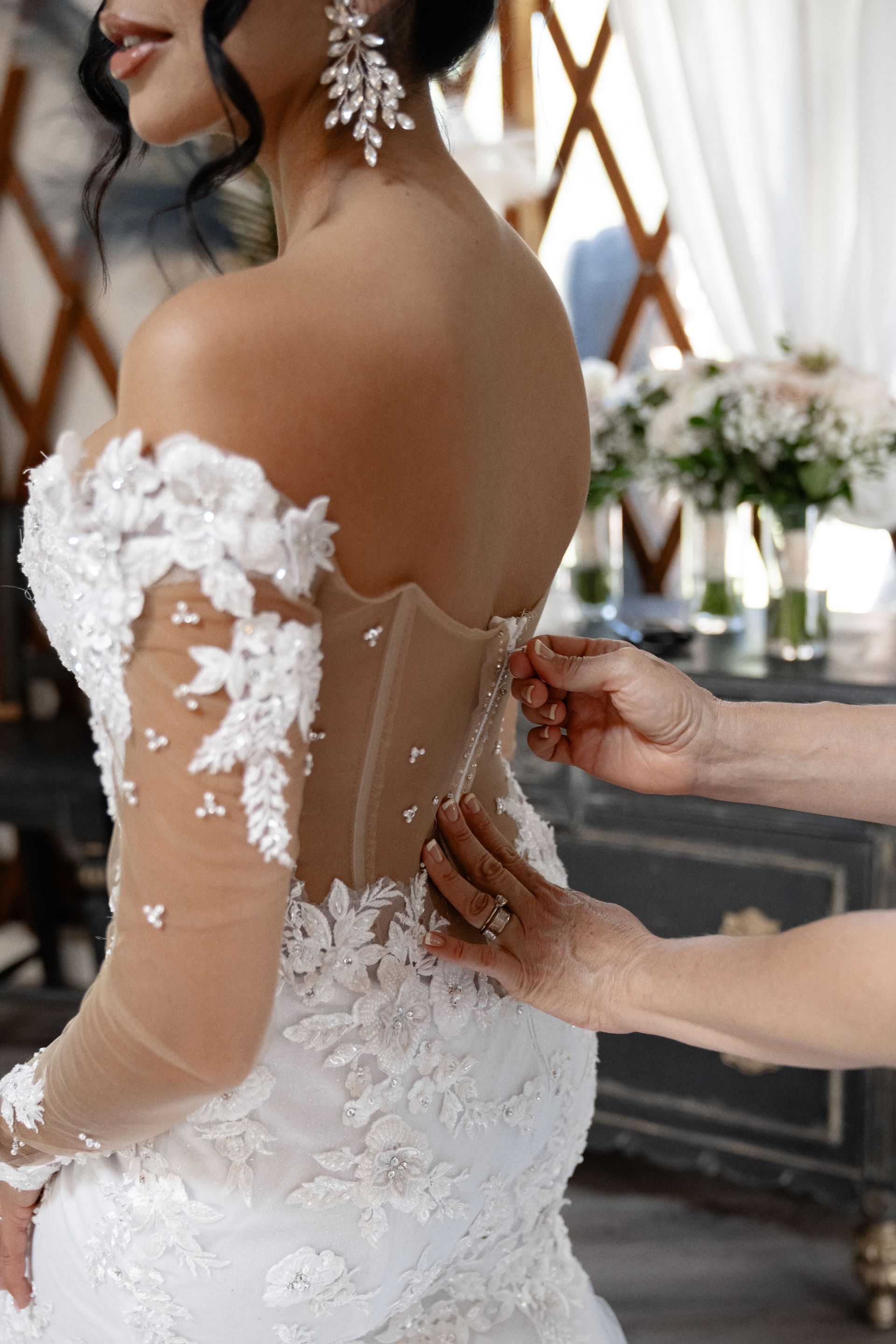 Bride in a white lace gown being fastened by hands, with floral decor in the background.