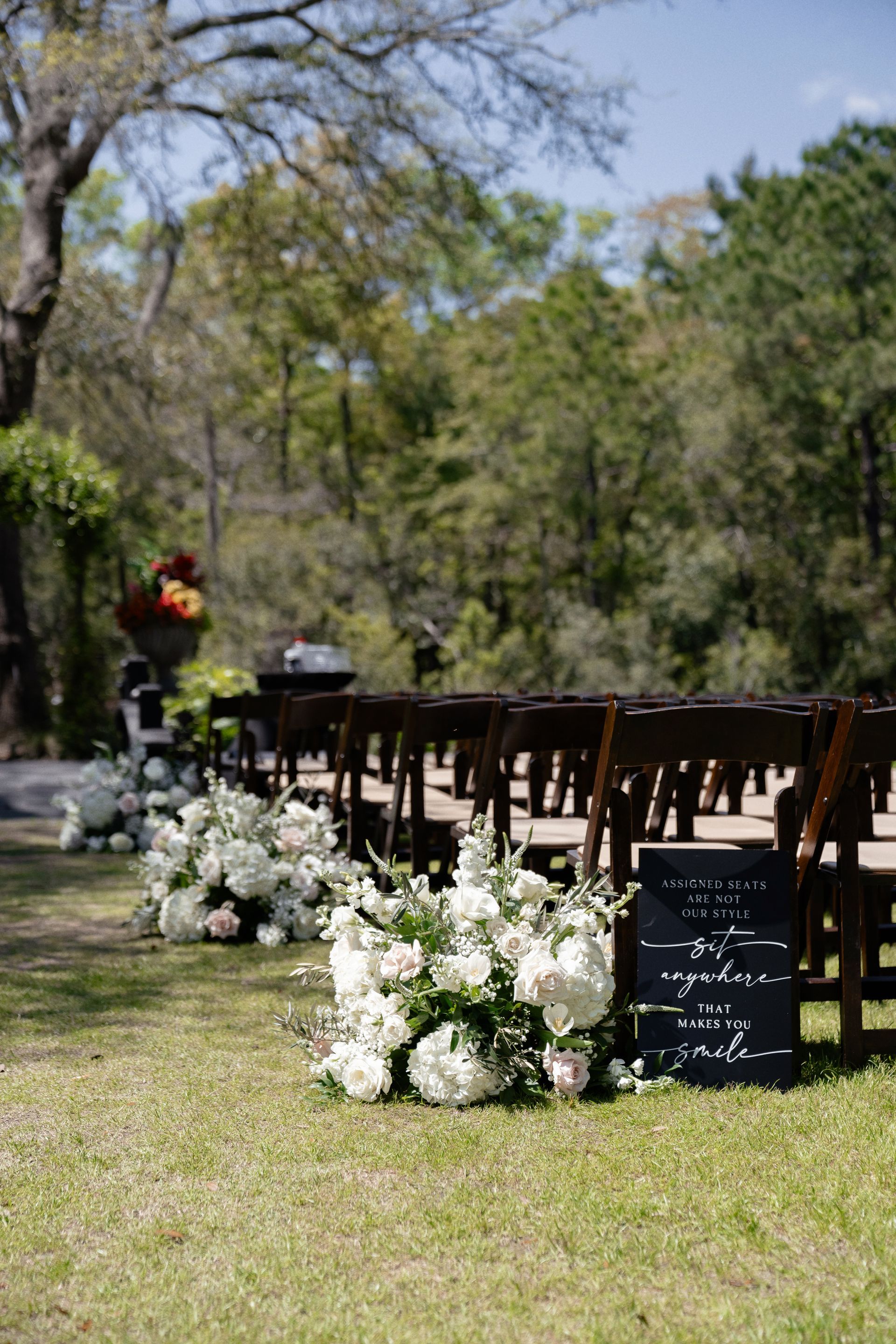 Outdoor wedding ceremony setup with wooden chairs, white floral arrangements, and a tree-lined lawn