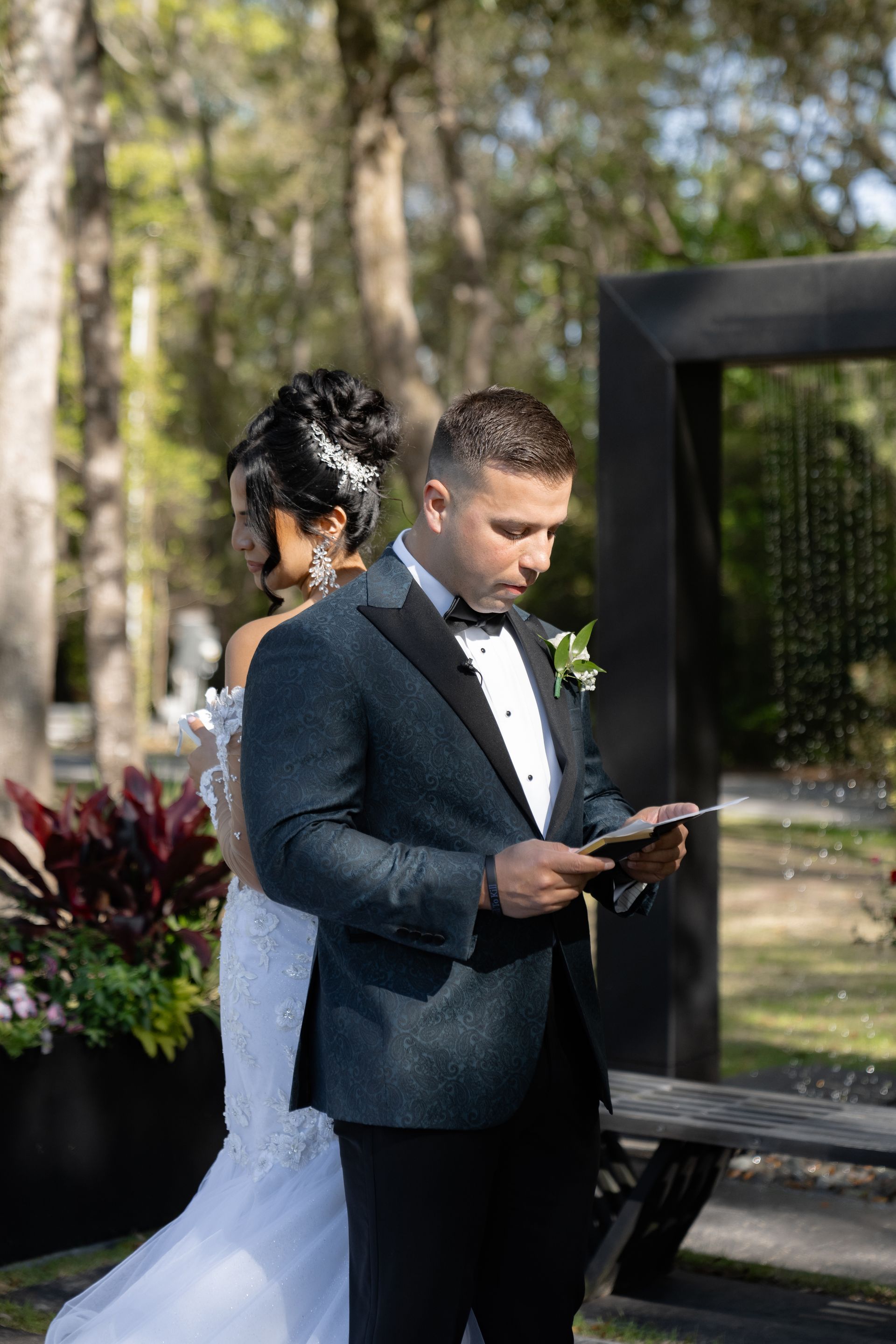 Man in a dark suit reading from a phone at an outdoor wedding ceremony, with a bride in a white dress behind him