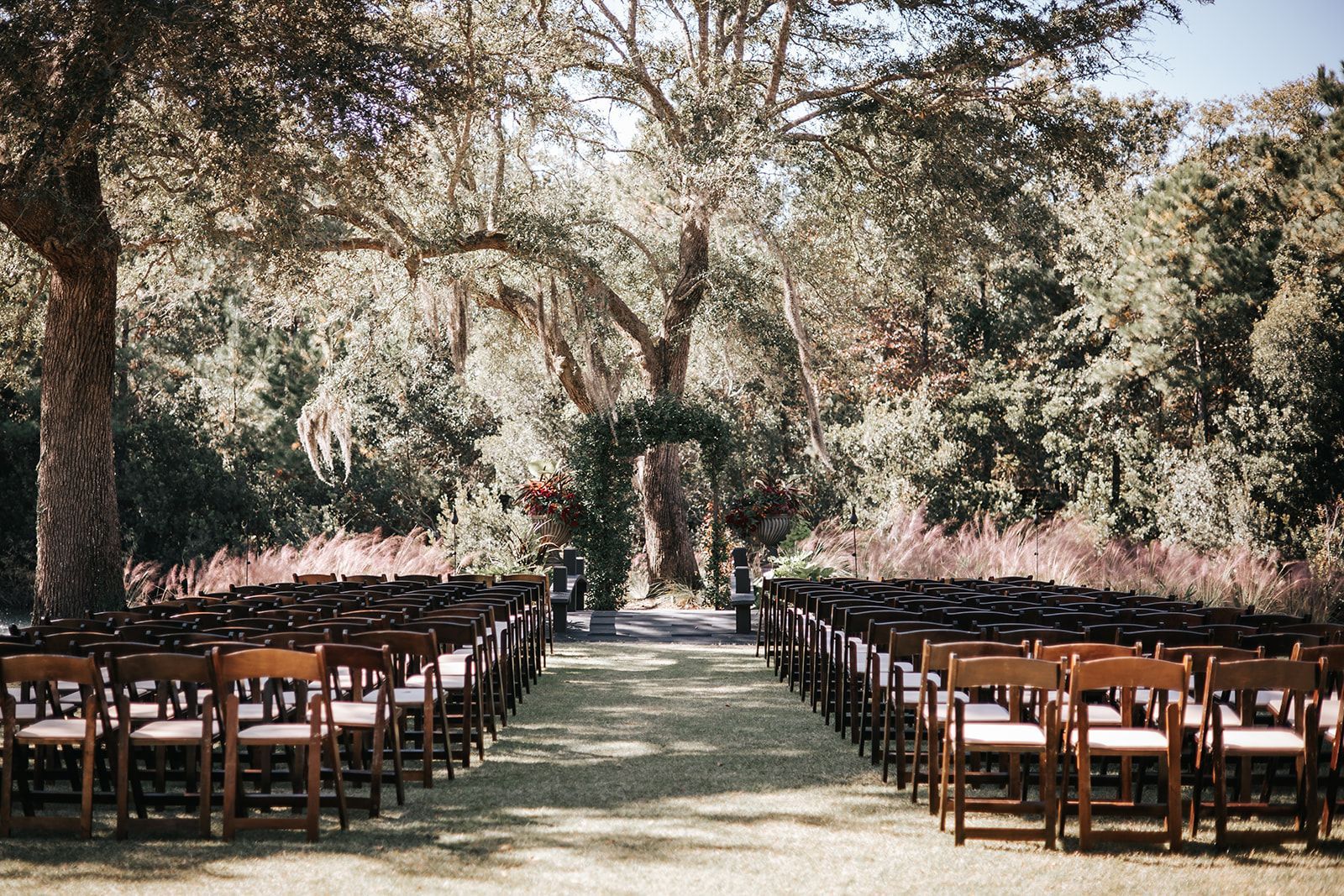 Outdoor wedding ceremony setup with rows of wooden chairs beneath trees lining a grassy aisle