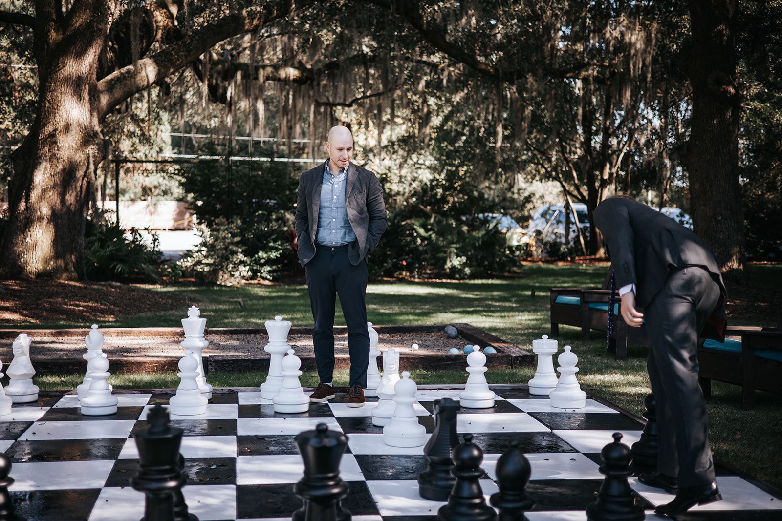 Two people stand on a giant outdoor chessboard in a park, one leaning over a chess piece.