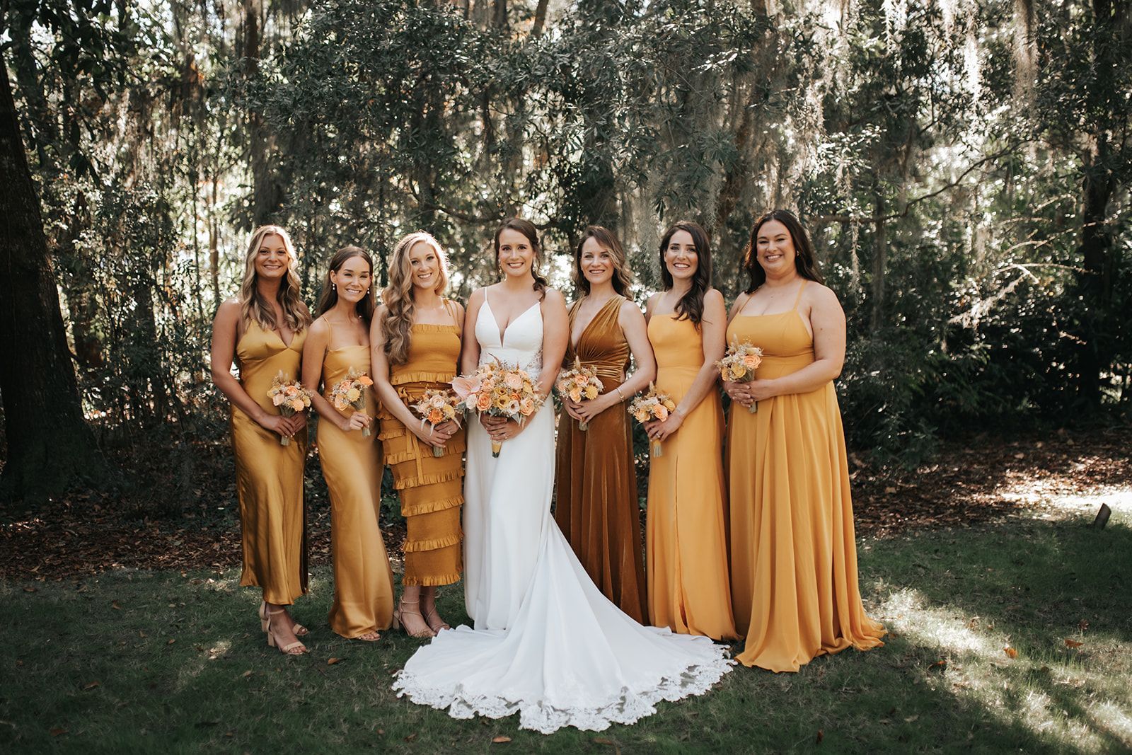 Bride and bridesmaids in gold dresses holding bouquets in a wooded outdoor setting