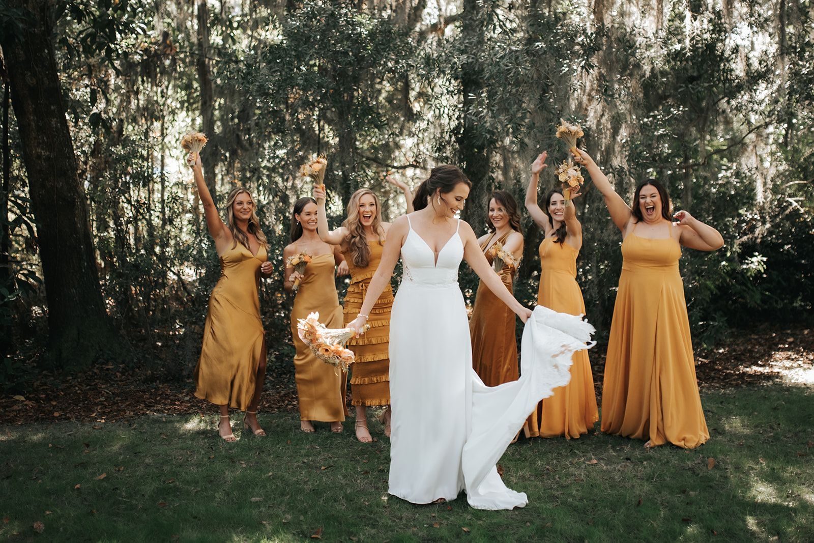 Bride and bridesmaids in gold dresses celebrating outdoors with raised arms and a white dog in front