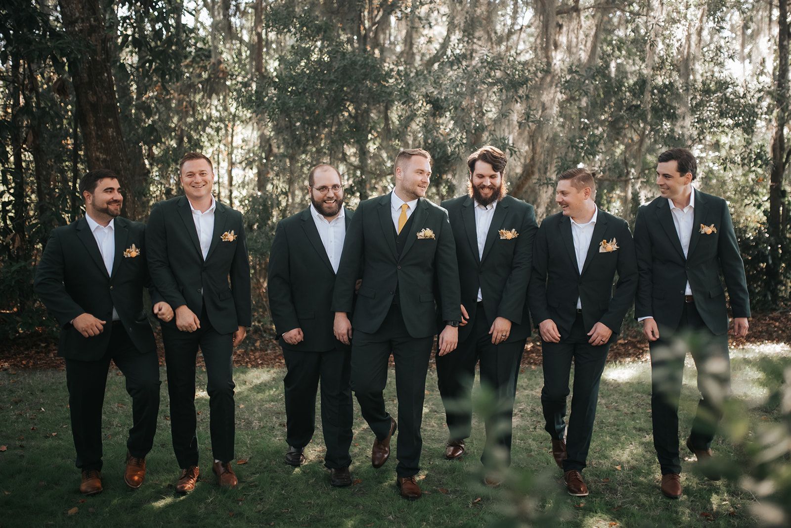 Six men in dark suits stand outdoors in a wooded area, posing for a formal group photo.