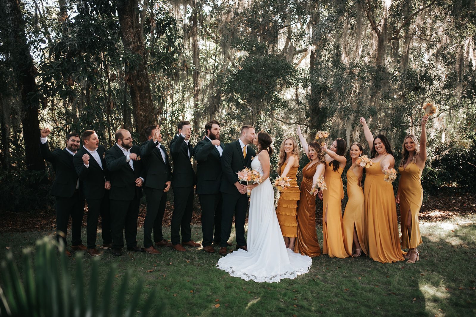 Wedding party posing outdoors beneath trees, bride in white dress with attendants in gold dresses.