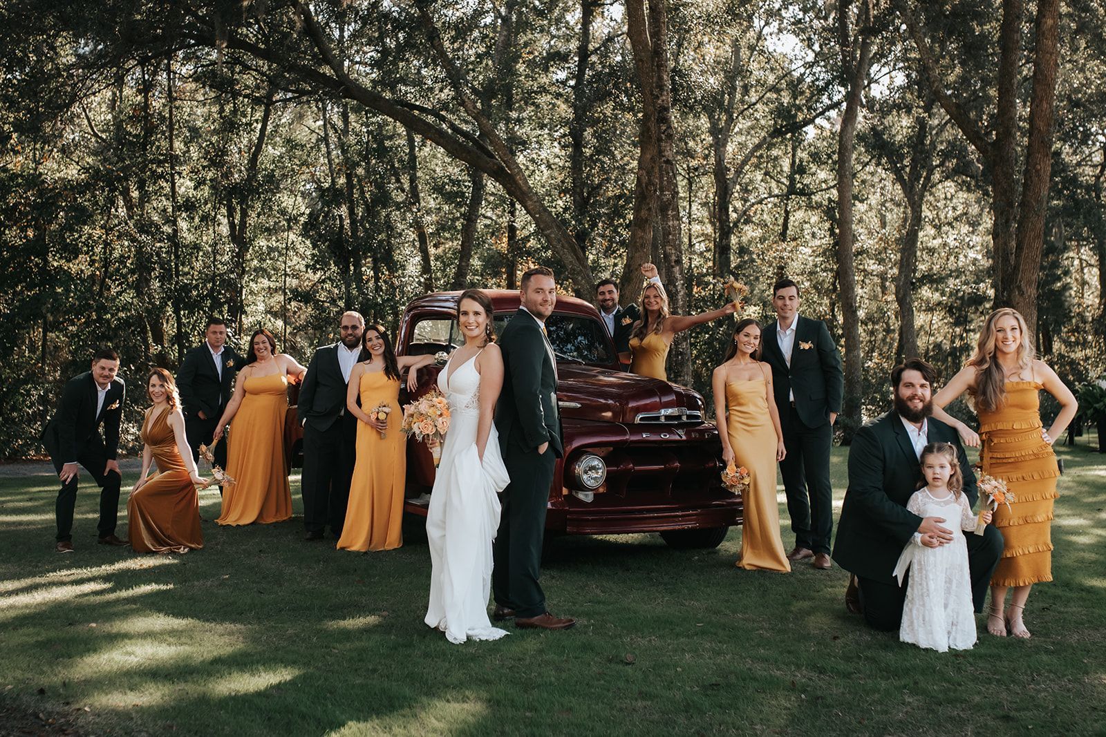 Wedding party posing outdoors around a dark red car under trees