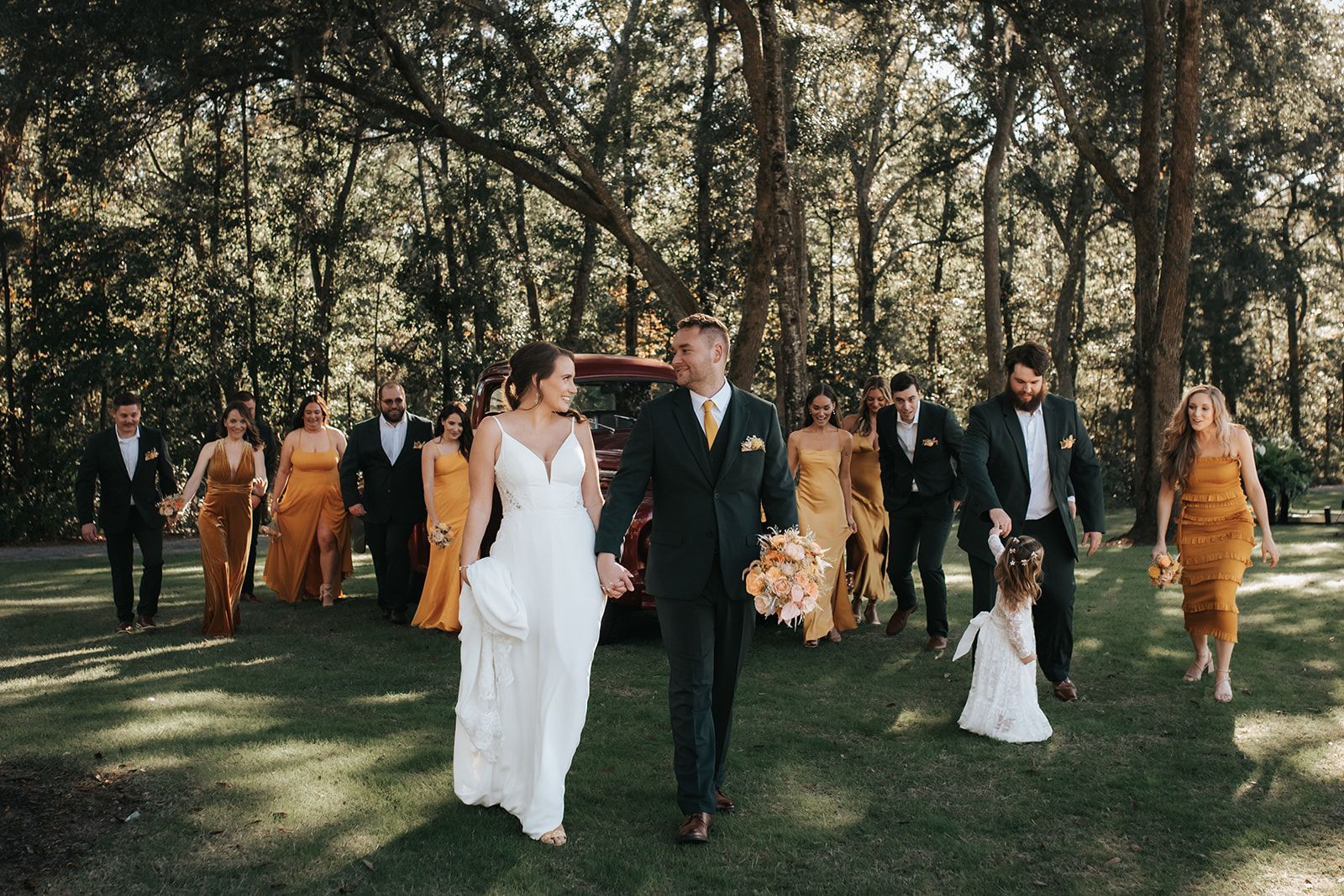 Wedding party walking outdoors under trees, with bride in white and groom in dark suit at center.