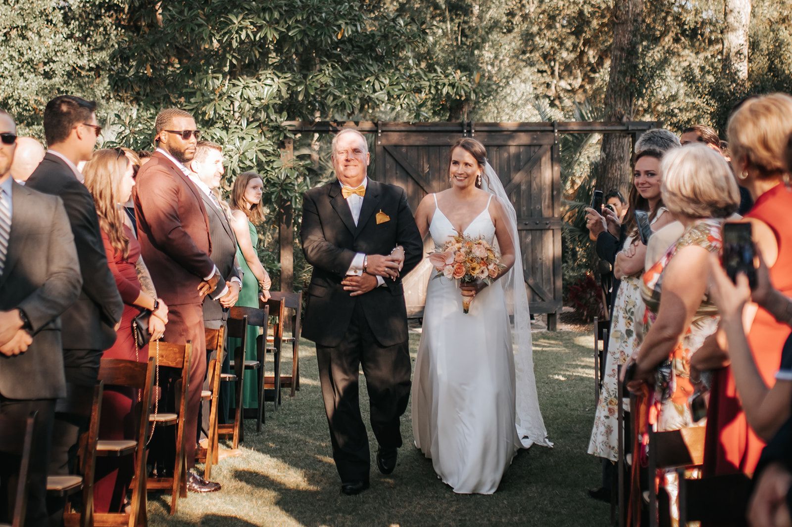 Bride and groom walk down an outdoor aisle between seated guests, with the bride holding a bouquet.