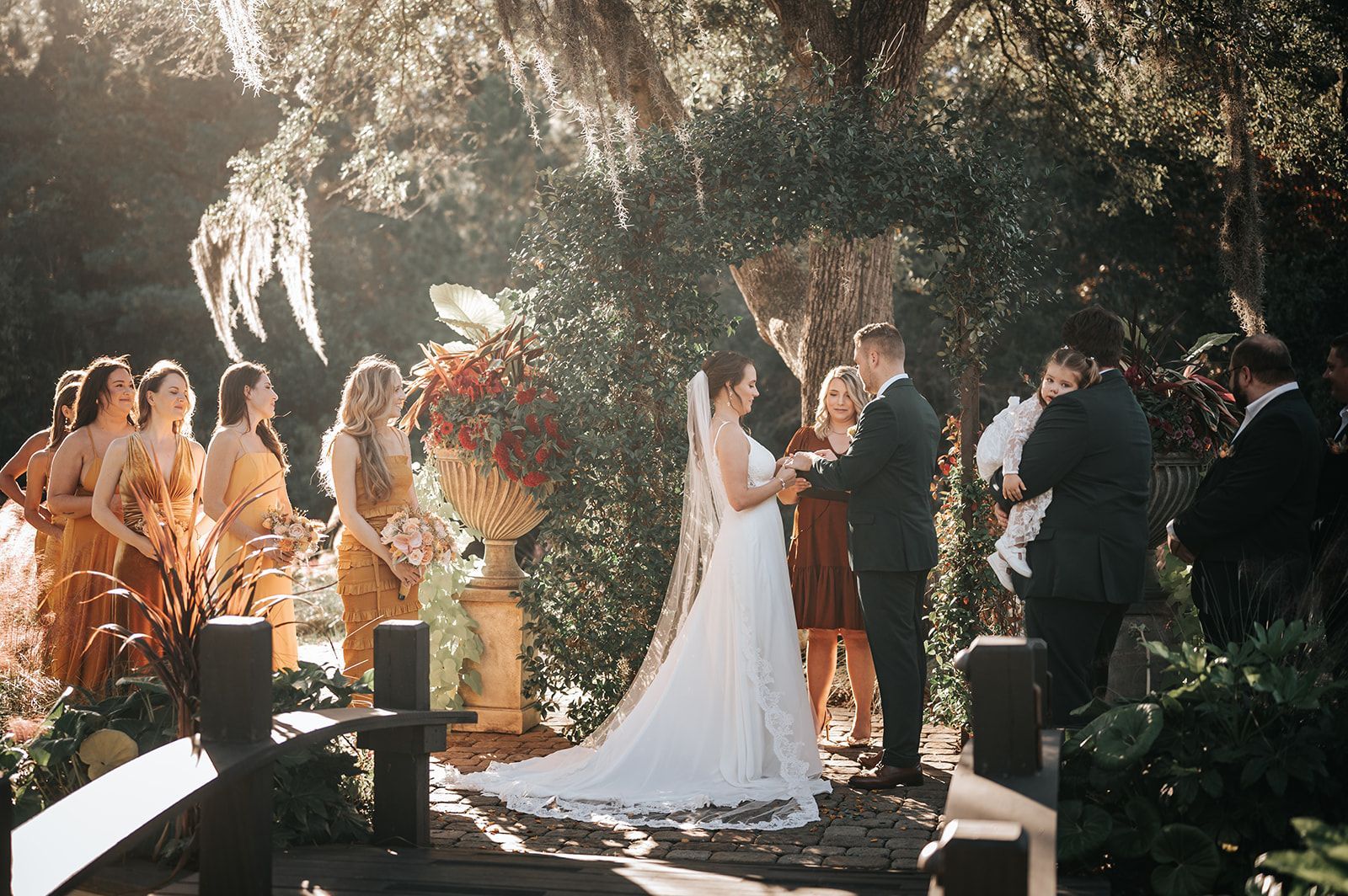 Outdoor wedding ceremony under trees, bride and groom exchanging vows with guests watching