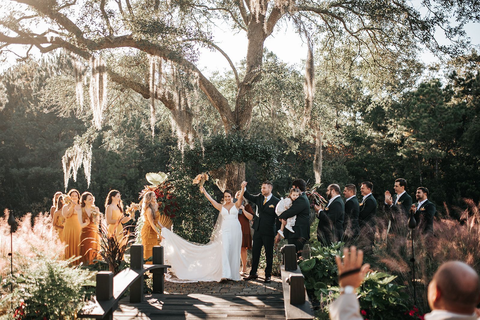 Outdoor wedding ceremony with bride and groom under trees, wedding party and guests gathered around