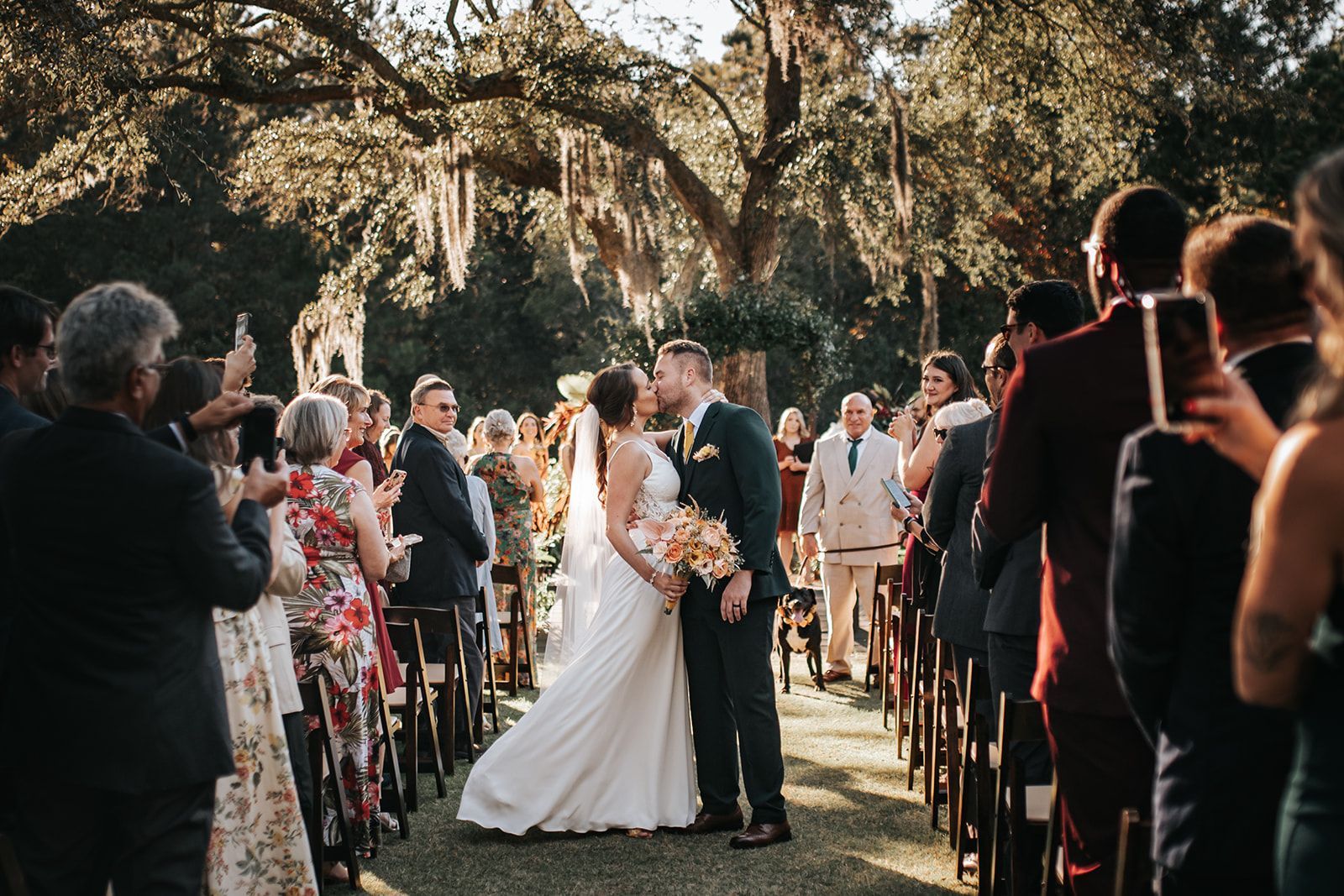 Bride and groom kissing under trees at an outdoor wedding ceremony, guests lining the aisle