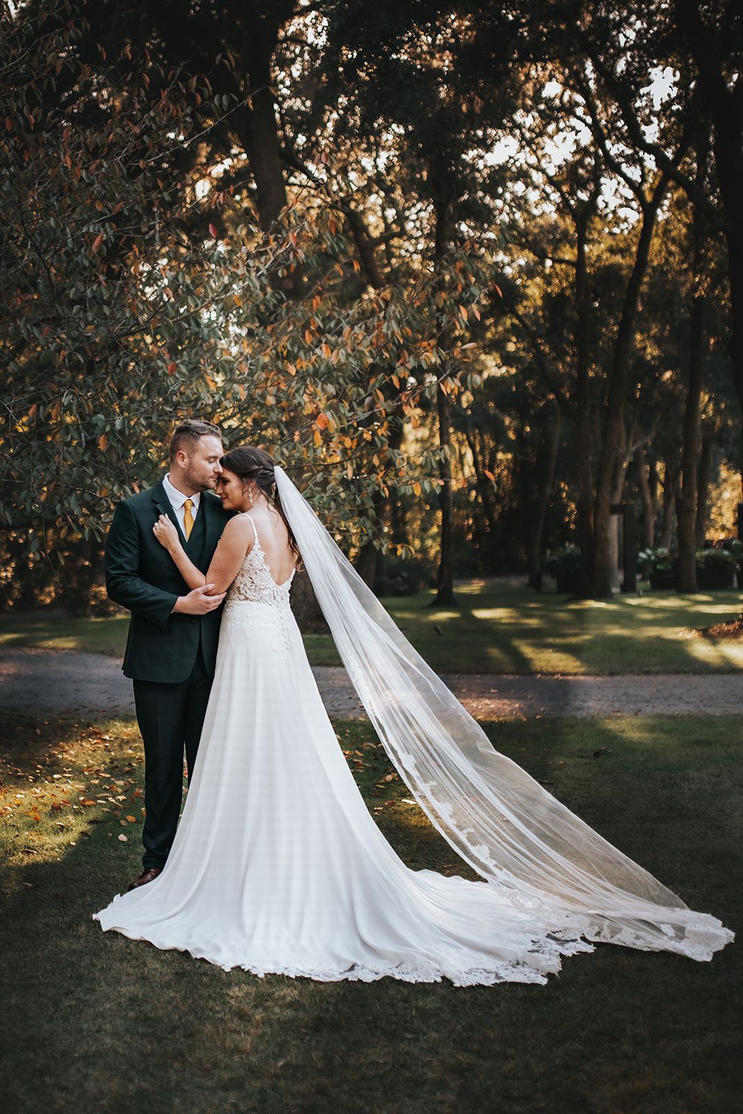 Bride and groom posing in a wooded park, her long veil trailing behind her dress