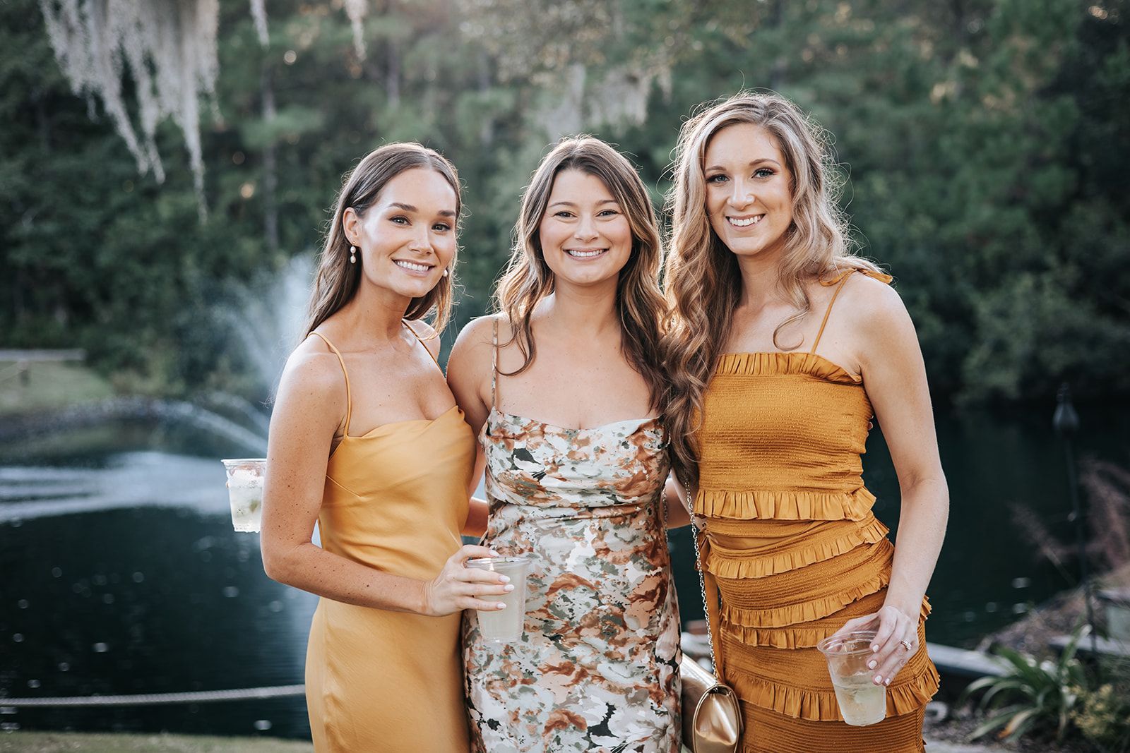 Three women in dresses posing outdoors by a river, smiling at the camera.