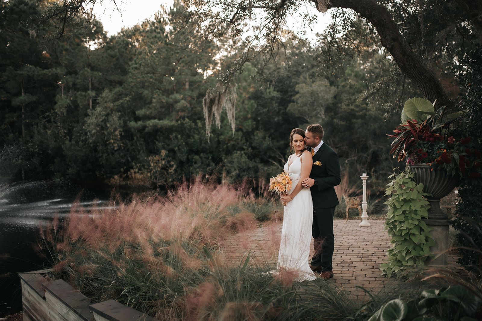 Couple in wedding attire standing in a garden, holding a bouquet amid tall grasses and trees.