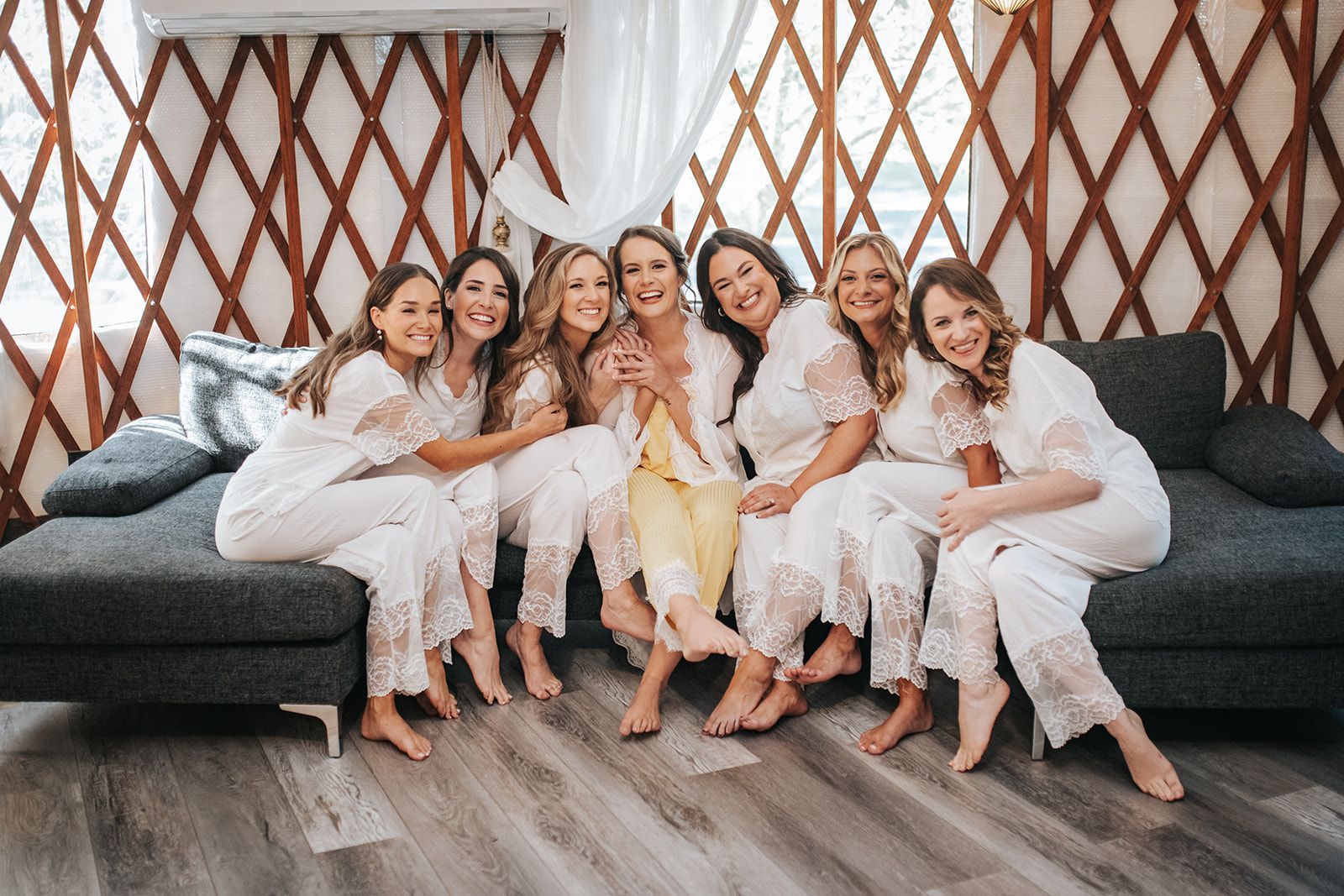 Group of six women in white outfits sitting on a gray couch in a decorated room with patterned walls