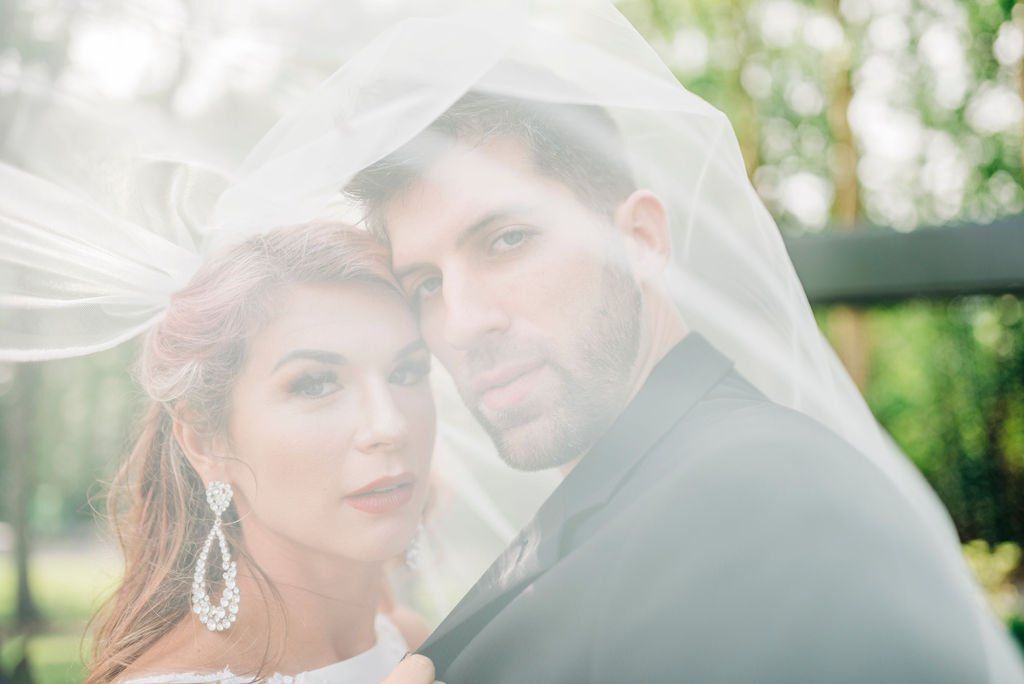 Bride and groom under a white veil outdoors, softly lit and embracing