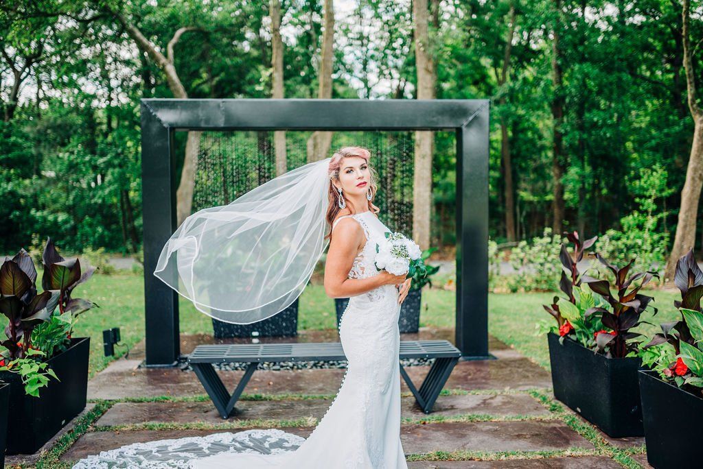 Bride in white dress and veil standing before a black frame in a lush garden, holding a bouquet.