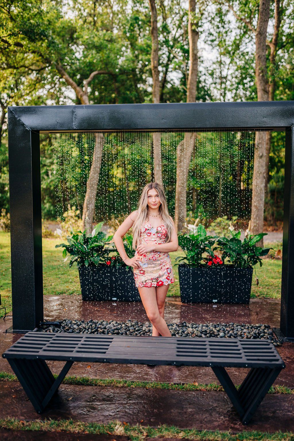 Person posing inside a black outdoor frame bench in a leafy park, with potted plants and trees behind.