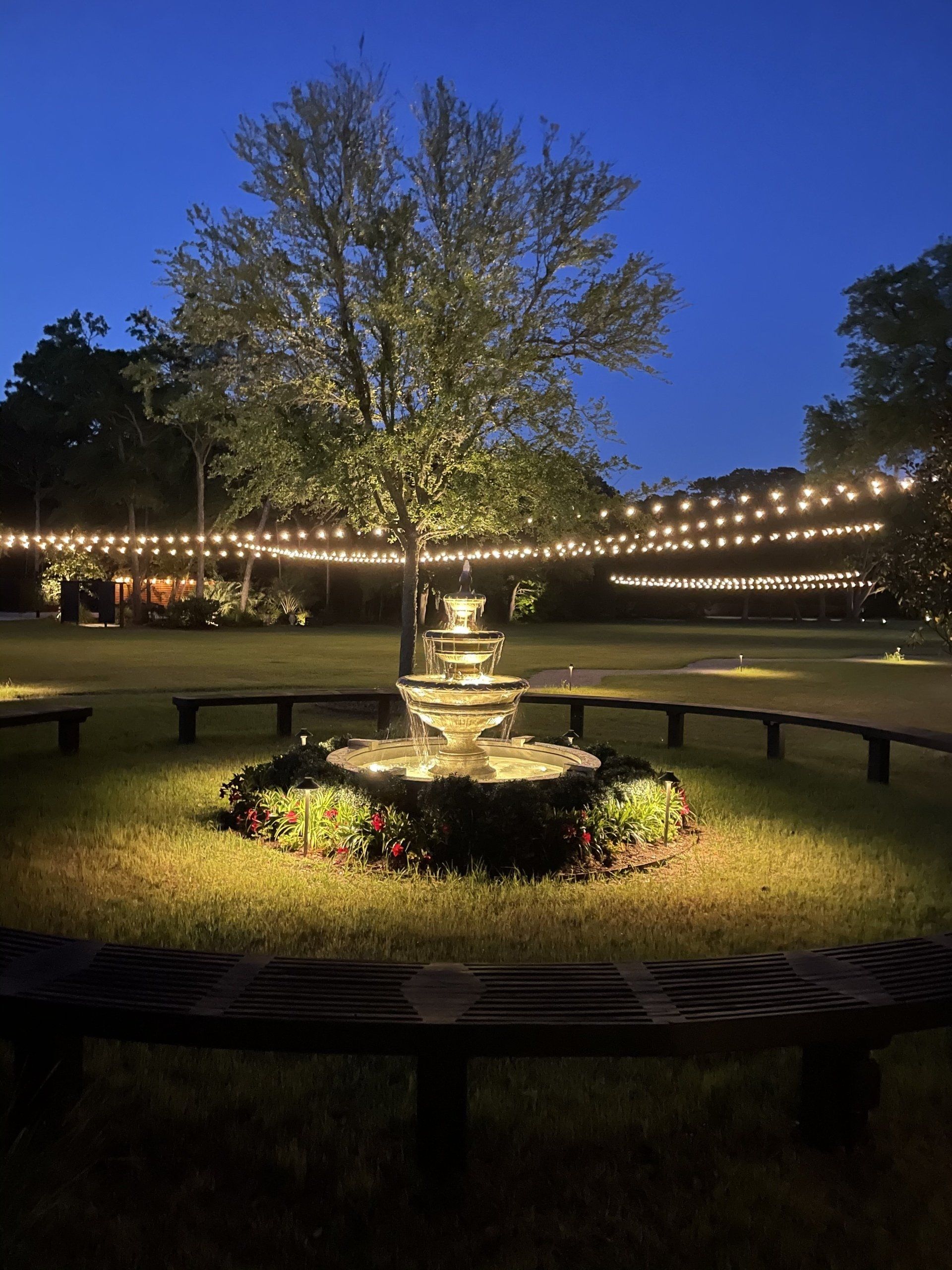 Lit garden fountain at dusk with string lights, benches, and trees in a park