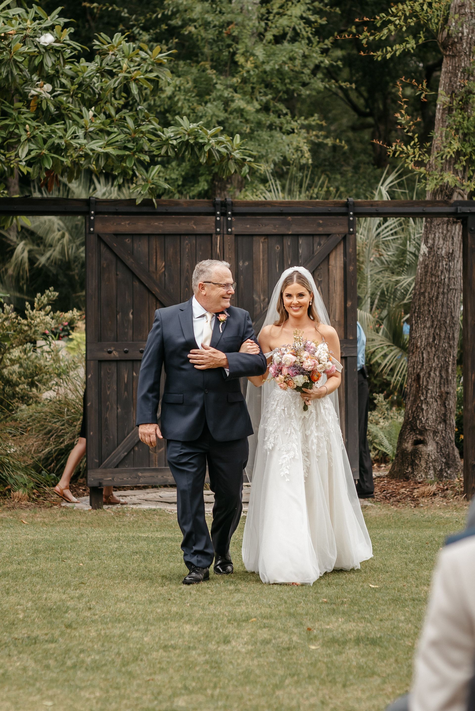 Bride in white gown walks arm-in-arm with man in dark suit in a garden wedding setting.