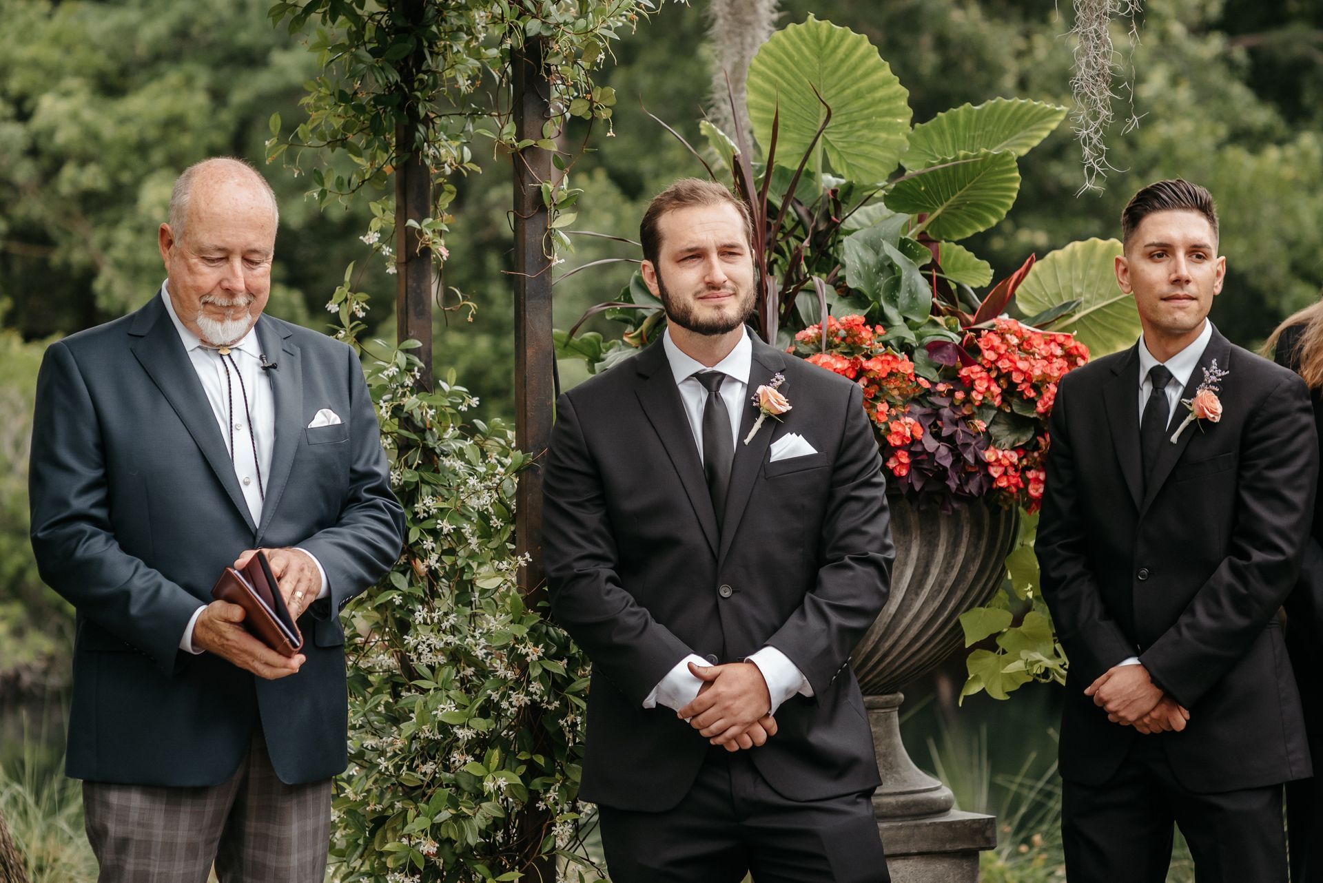 Three men in suits stand outdoors beside a floral arrangement at a formal event.