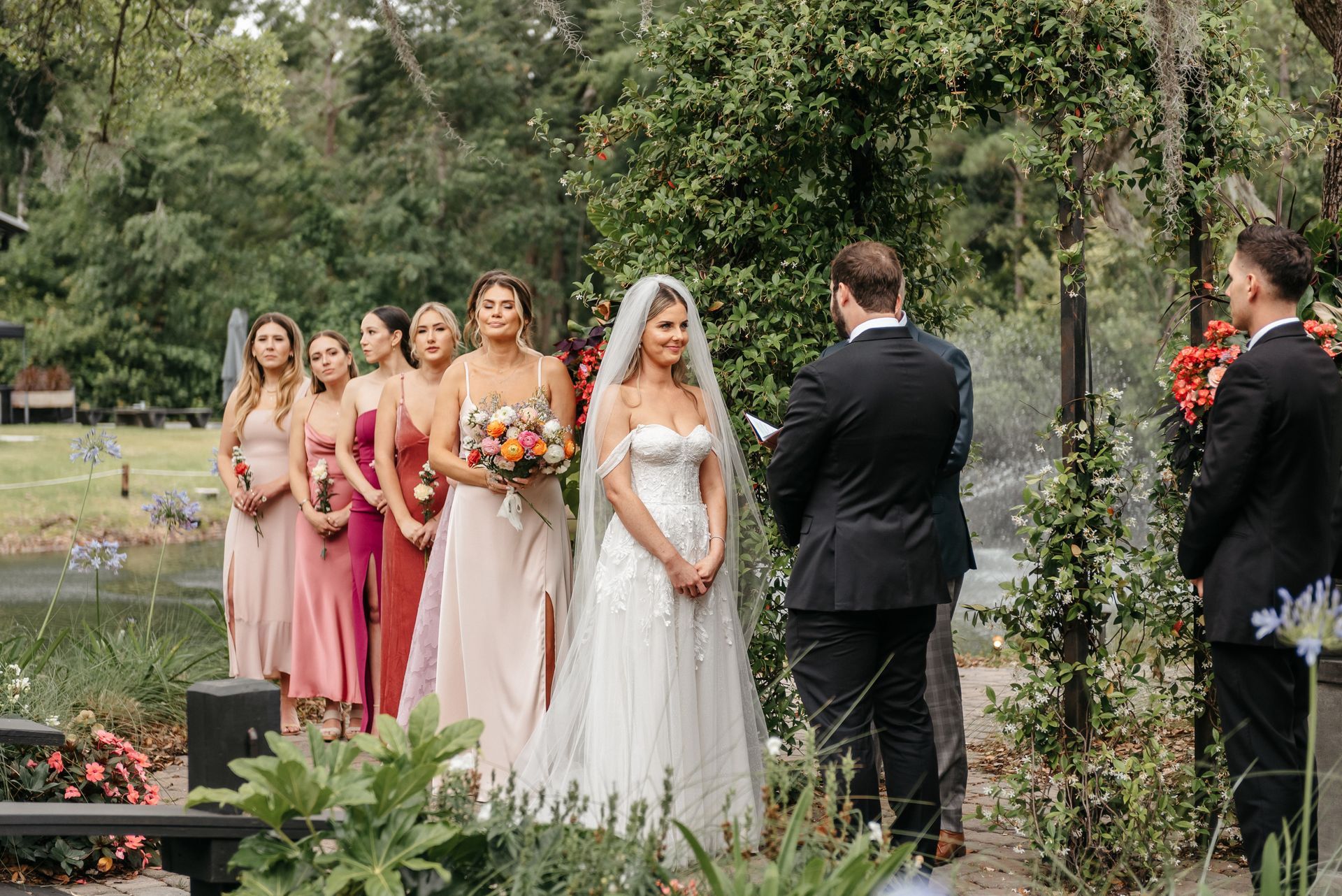 Outdoor wedding ceremony with bride and groom facing each other, bridesmaids in pastel dresses nearby