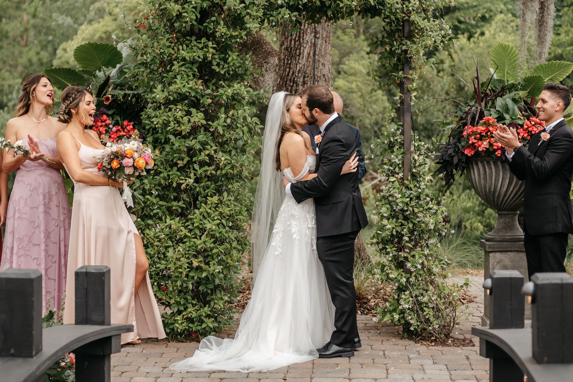 Wedding couple kissing at an outdoor ceremony, with bridesmaids holding bouquets nearby