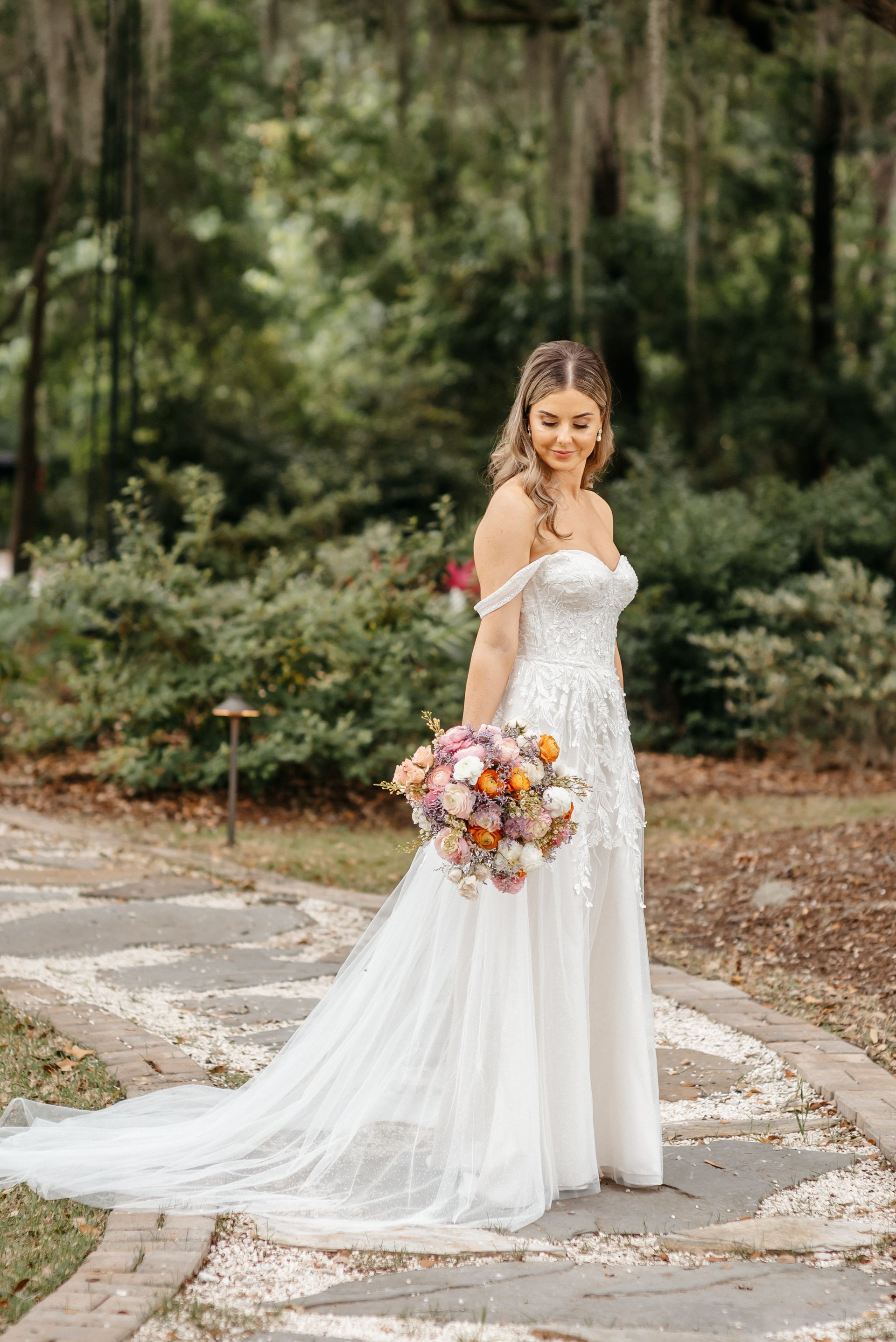 Bride in a white gown holding a colorful bouquet on a garden path, smiling over her shoulder.