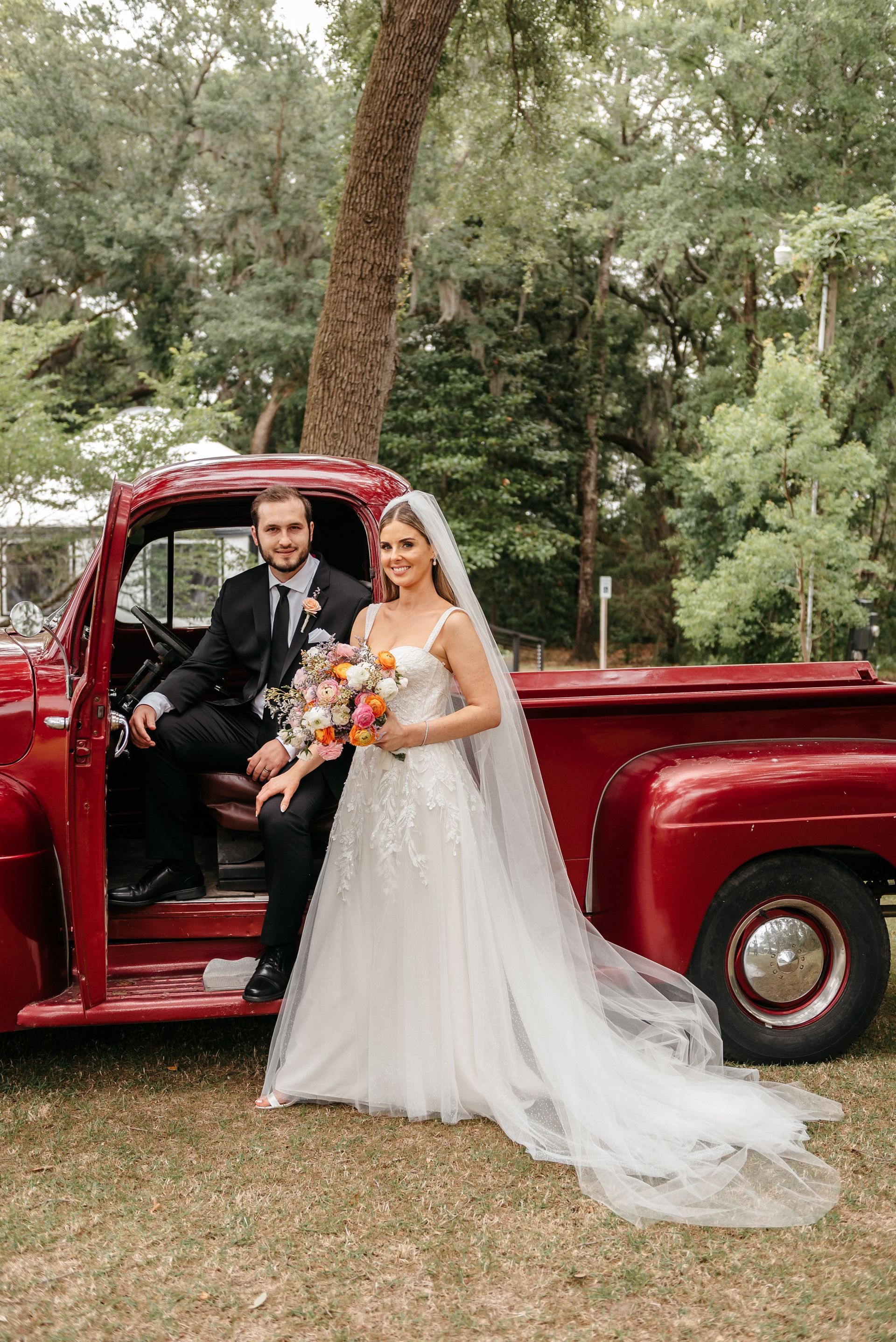 Bride and groom standing by a red vintage pickup truck in a wooded outdoor setting
