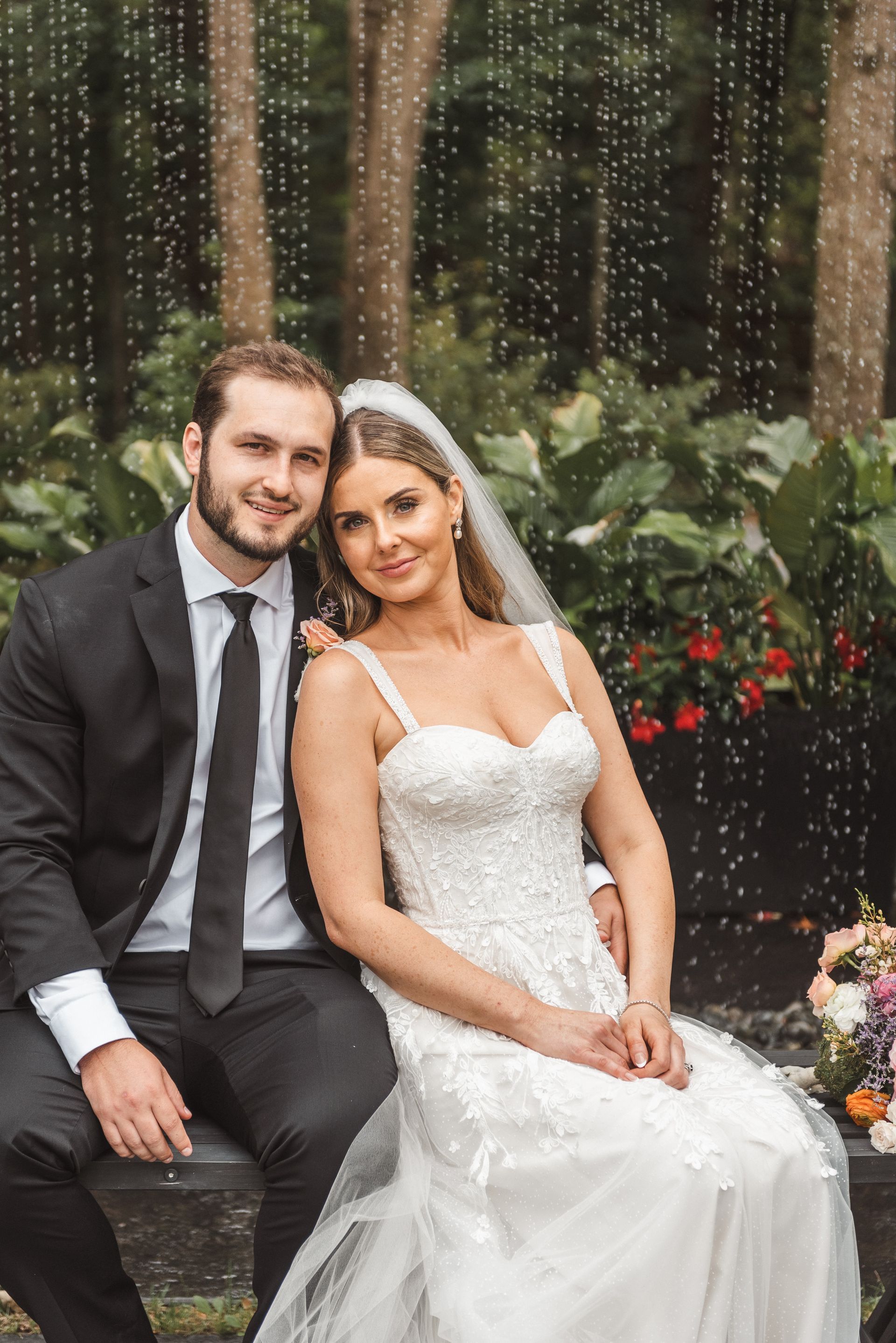 Bride and groom seated outdoors in wedding attire, smiling beside flowers and greenery