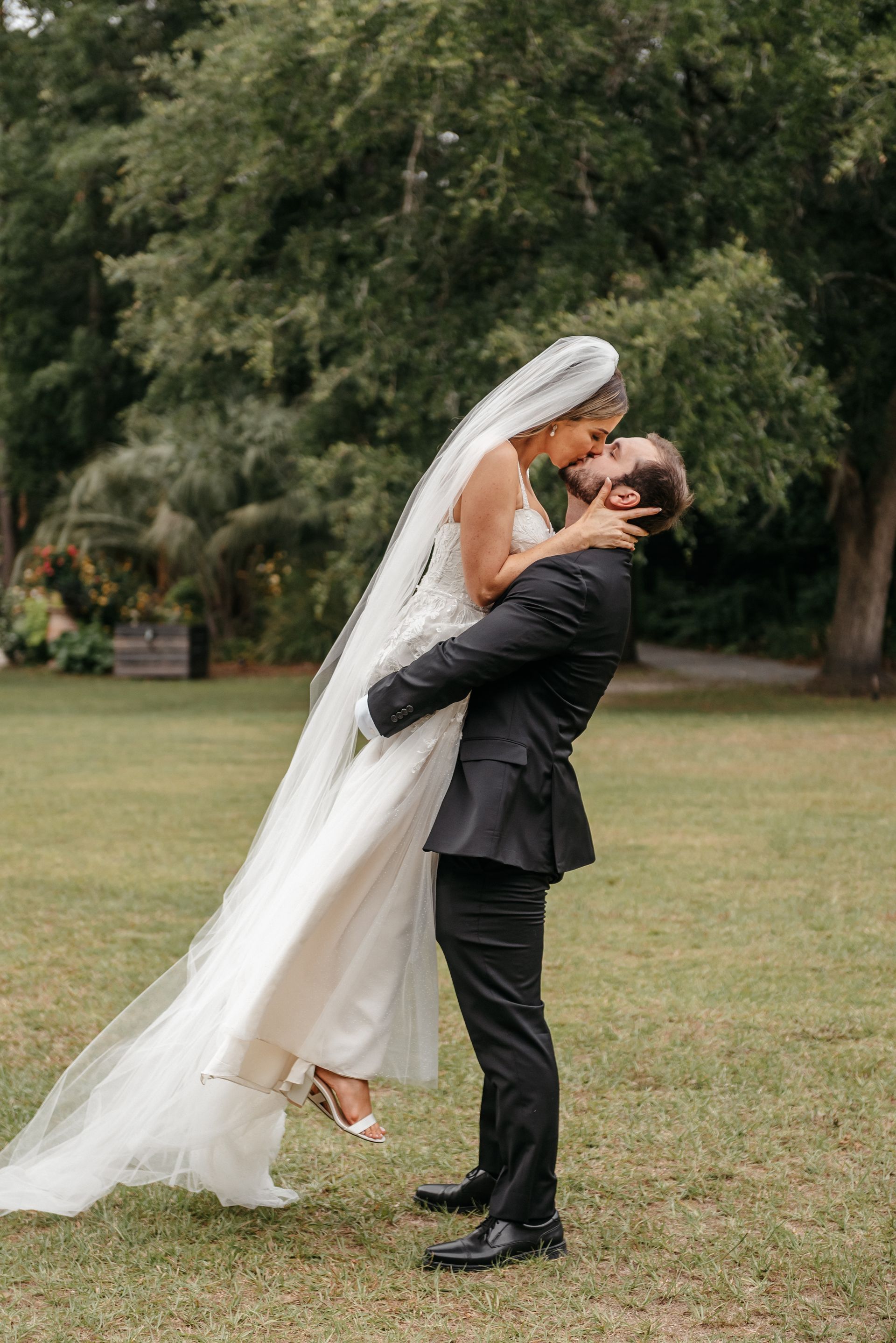 Bride and groom in wedding attire sharing a kiss in a grassy park with trees behind them