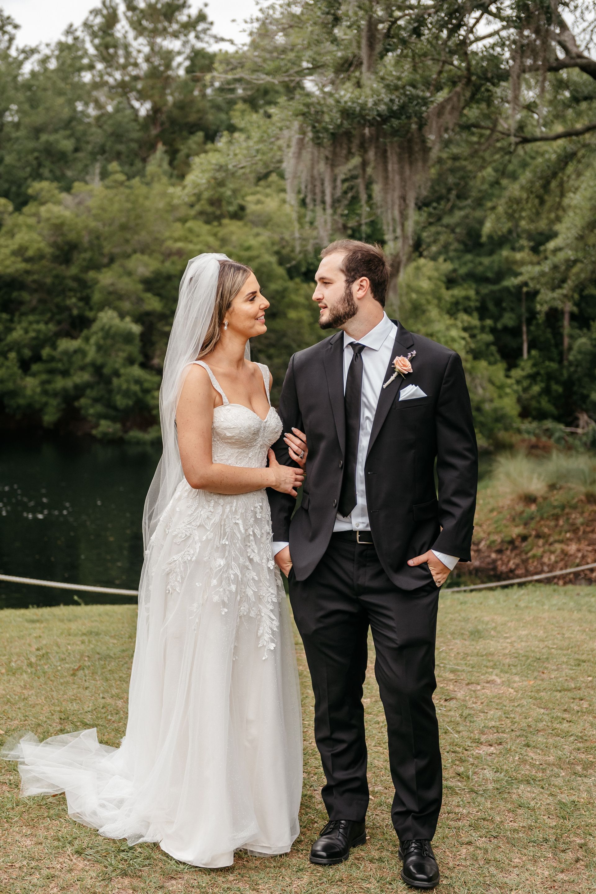 Bride and groom in wedding attire standing by a lake, holding hands and smiling at each other.